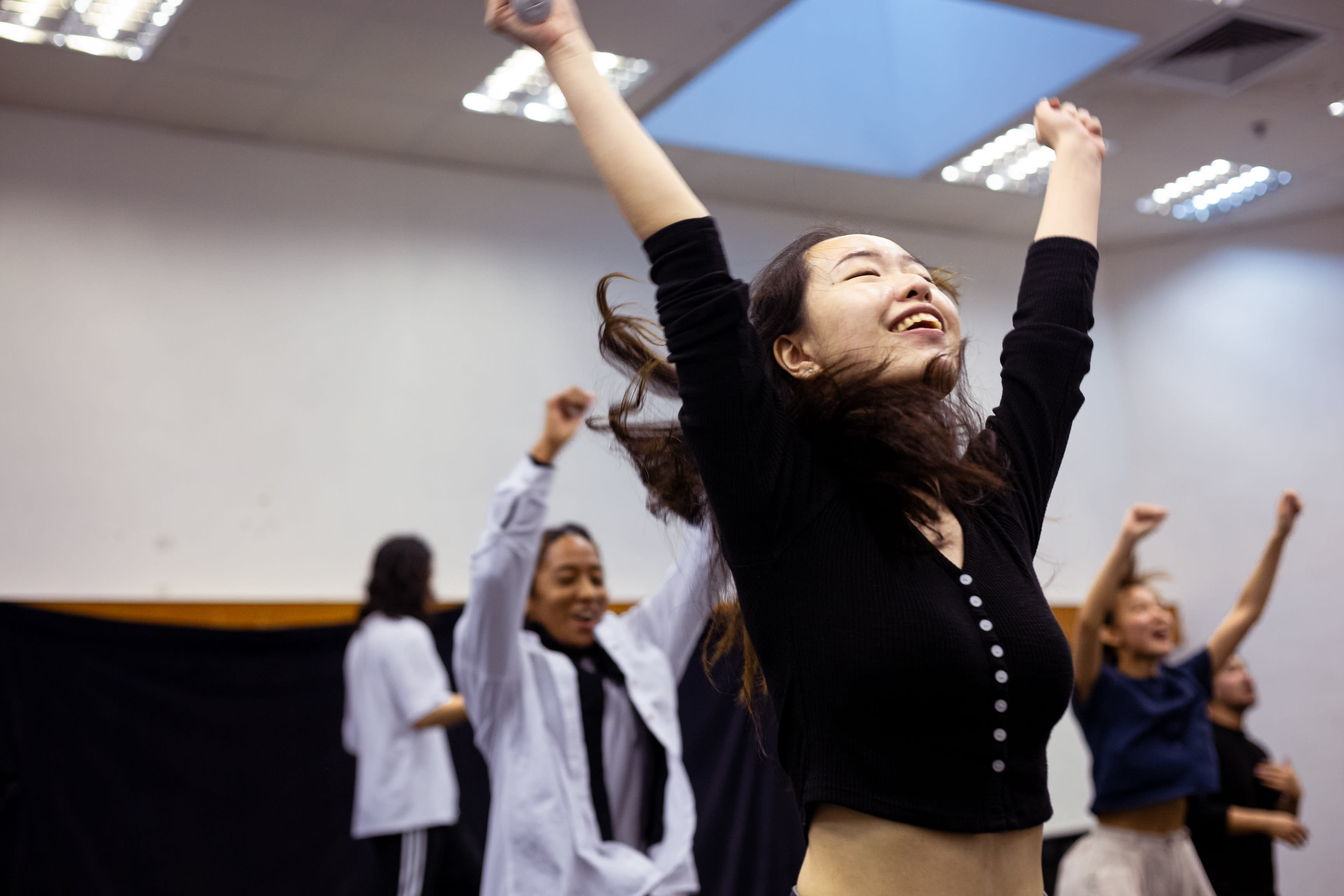 In a rehearsal for the contemporary Hong Kong theatre production 'Songs of Innocence and Experience' by Rooftop Productions, a performer playing Abdiel (Sum Cheuk-yiu) stands in the foreground, eyes closed and head tilted back, arms raised high in an expression of pure, ecstatic triumph. Behind her, other cast members, including Caroline Chan, are captured in motion, their arms raised as they participate in the intense, physical storytelling of this experimental stage performance. Songs of Innocence and Experience Rehearsal