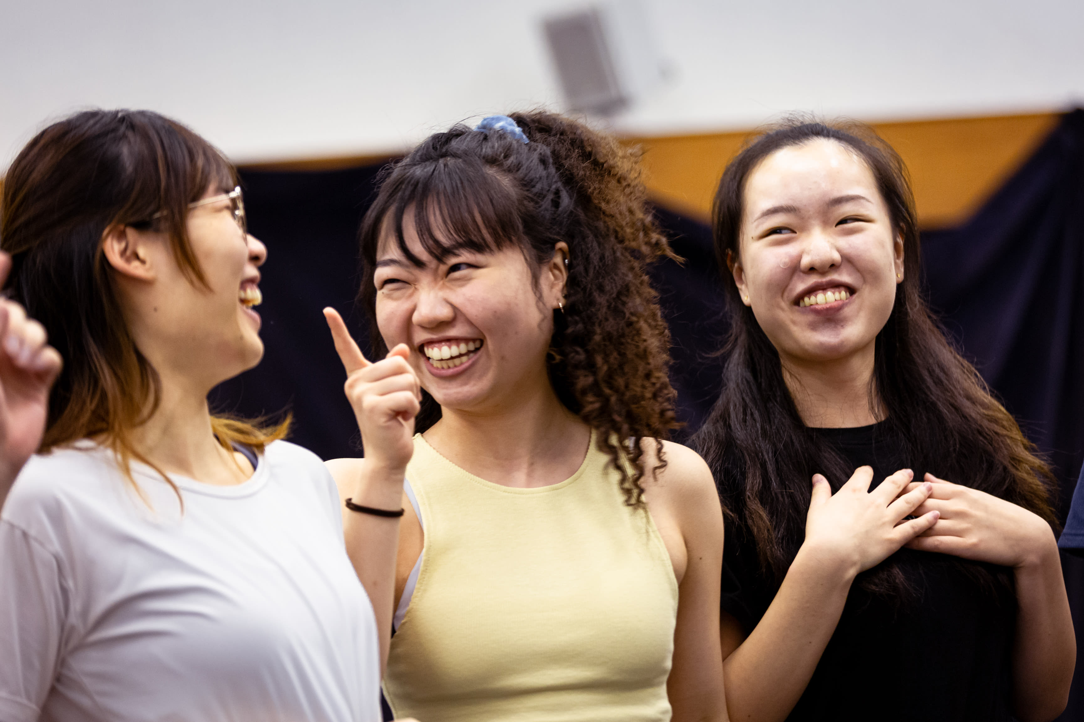 Three performers—Lee On-sang, Sum Cheuk-yiu, and Lau Ka-ying—share a lighthearted moment during a rehearsal for the Rooftop Productions contemporary theatre piece, 'Songs of Innocence and Experience'. Sum Cheuk-yiu (playing Abdiel) points playfully while laughing, flanked by Lee On-sang (playing Moloch) and Lau Ka-ying (playing Belial), who expresses heartfelt joy with her hands over her chest. The scene captures the collaborative spirit of HK stage performance. Songs of Innocence and Experience Rehearsal.