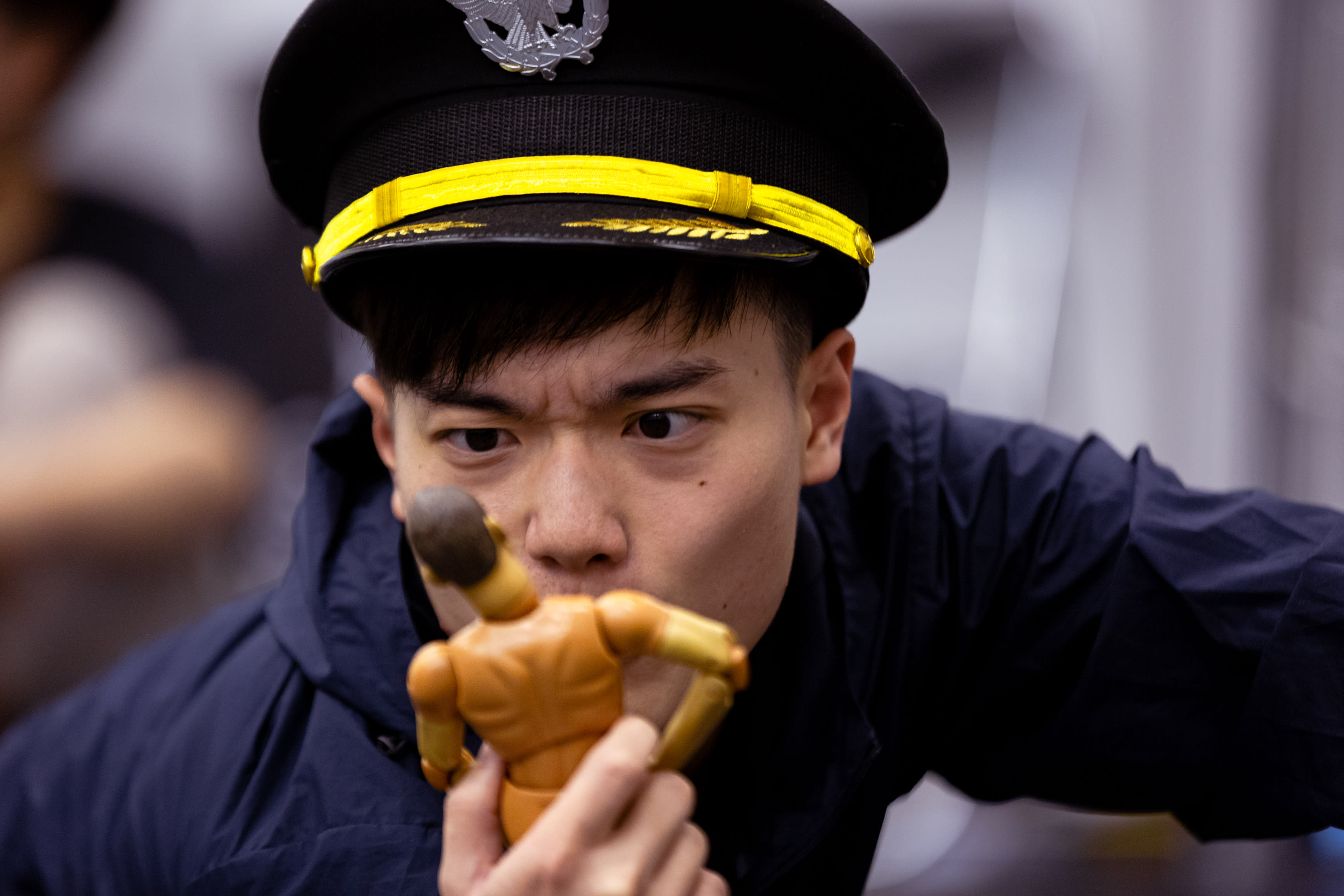 Close-up of actor Lo Man-chak in a rehearsal for Rooftop Productions' contemporary theatre piece, Songs of Innocence and Experience. Wearing a dark police-style cap with a gold band, he holds a small, featureless wooden mannequin figure near his face, his expression intense and focused as he contemplates the figurine. The atmospheric lighting captures the raw, experimental nature of Hong Kong stage performance. Songs of Innocence and Experience Rehearsal.