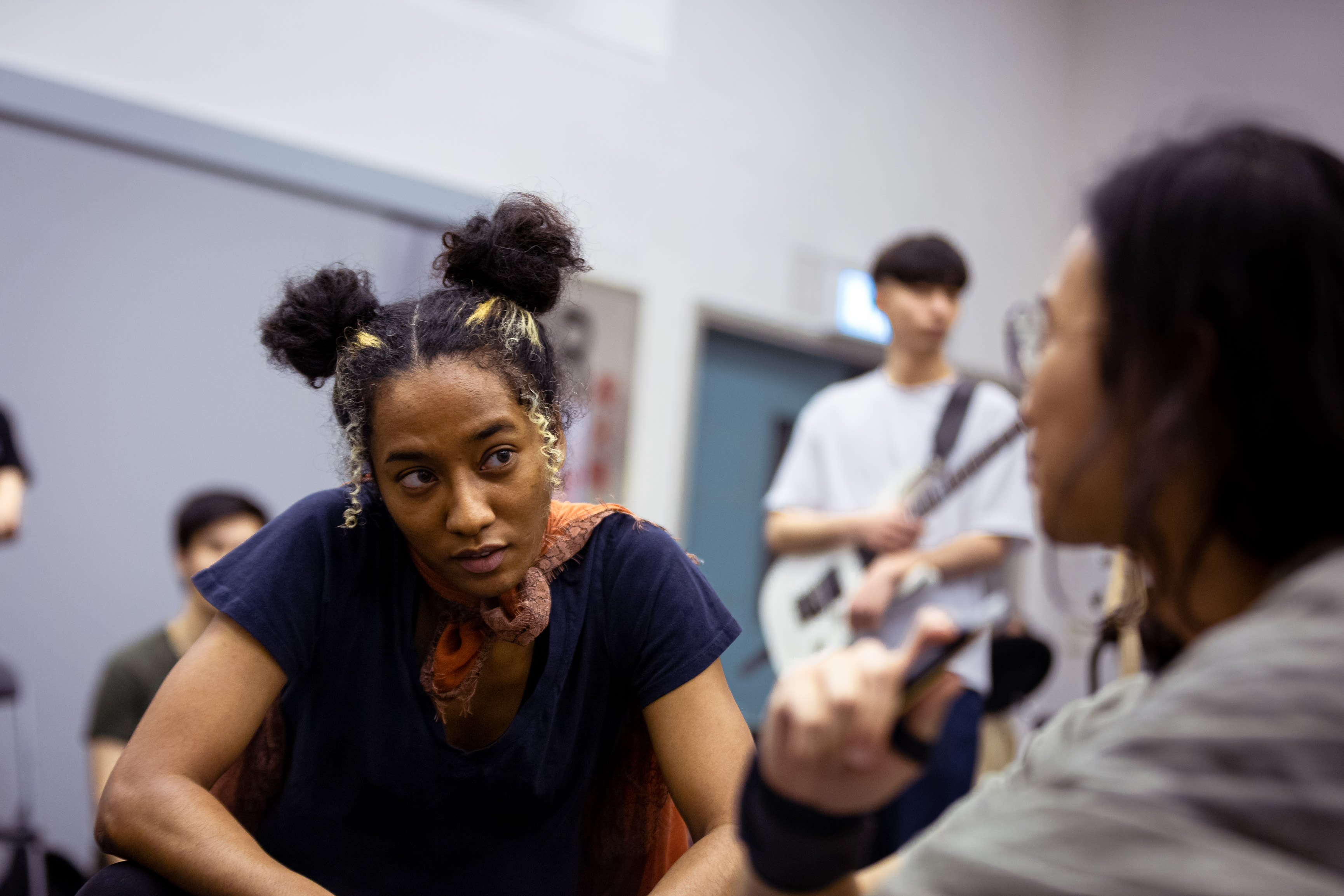 In a candid rehearsal for Rooftop Productions' contemporary theatre piece 'Songs of Innocence and Experience', actress Caroline Chan (cast as Satan/Lucifer) leans forward with an intense, focused gaze during a discussion. She wears a dark t-shirt and an orange scarf, her hair styled in two buns. In the background, another performer with a guitar is slightly out of focus, capturing the creative atmosphere of a Hong Kong stage production. Songs of Innocence and Experience Rehearsal.