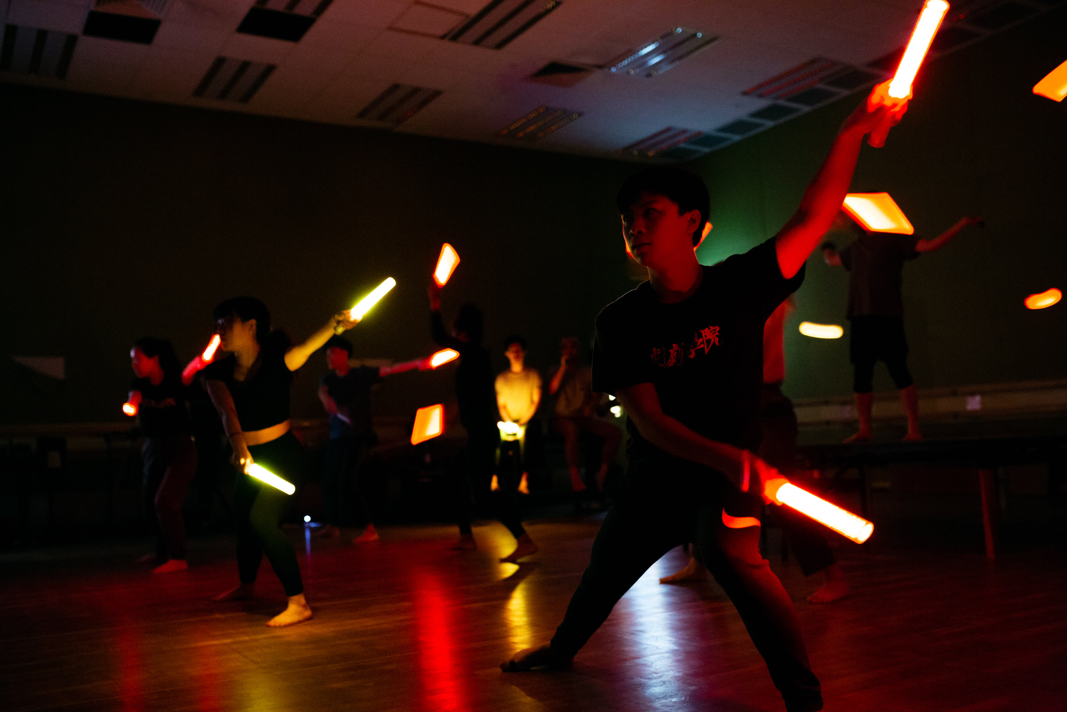 In a dim, atmospheric rehearsal room, performers from Rooftop Productions engage in an intense scene for their contemporary Hong Kong theatre piece, Songs of Innocence and Experience. Chew Yu-yeung, cast as Mammon, stands in the foreground with a focused, dynamic stance, wielding a glowing orange light stick high in the air. Other ensemble members are positioned in the background, their movements synchronized with similar light sticks to create streaks of motion. The lighting design emphasizes the contrast between shadow and the vibrant orange glow, capturing the theatrical energy of the rehearsal. Songs of Innocence and Experience Rehearsal.