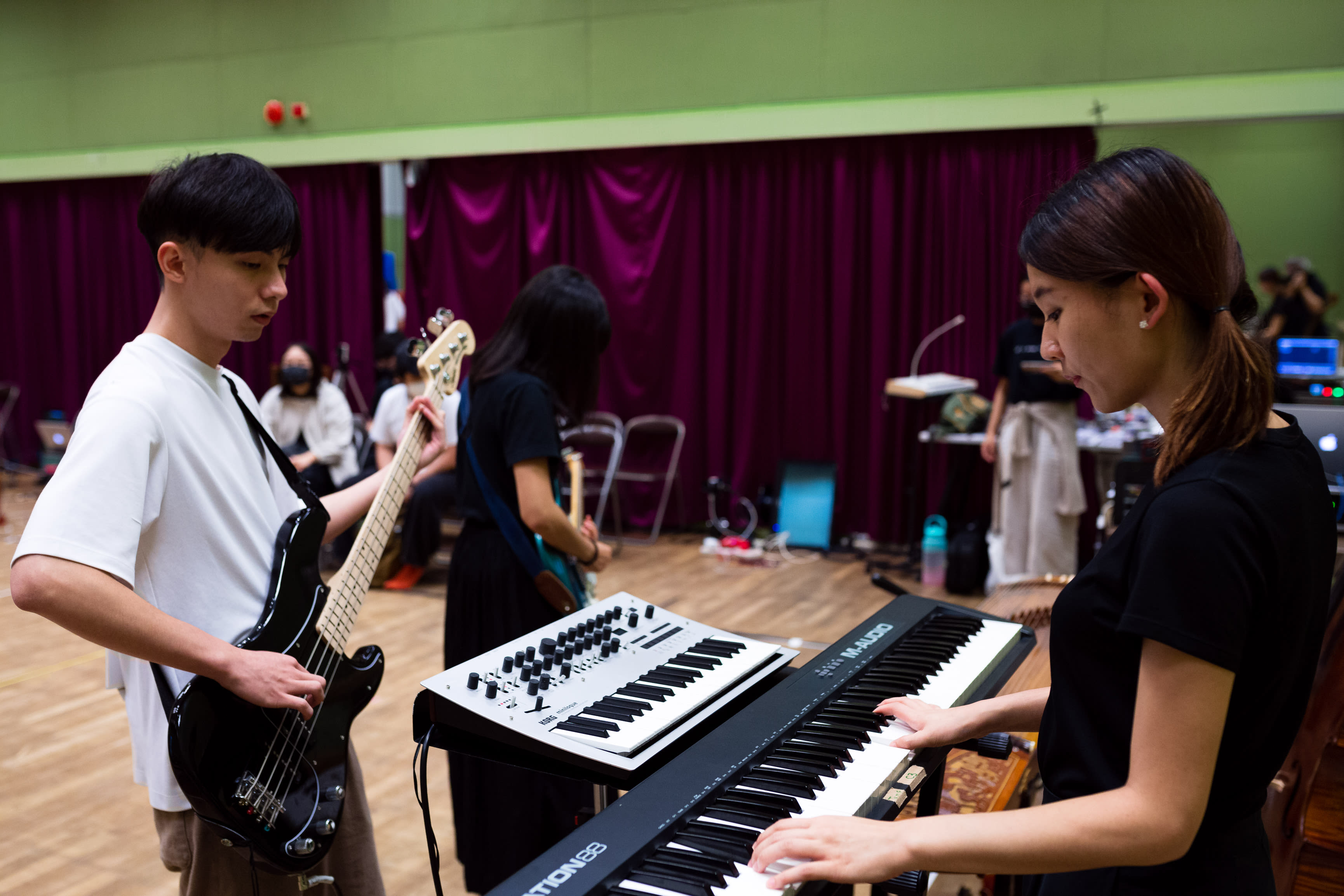 In a rehearsal for Rooftop Productions' contemporary theatre piece 'Songs of Innocence and Experience', band leader Wu Lui-fung stands left, focused on playing his black bass guitar. To the right, Tsang Yuen-lam Ivy plays a keyboard, her expression intent and concentrated. In the background, another performer works with a synthesizer in a dimly lit studio featuring dark stage curtains. This image captures the artistic rigor behind the scenes of a Hong Kong stage performance: 'Songs of Innocence and Experience Rehearsal.'