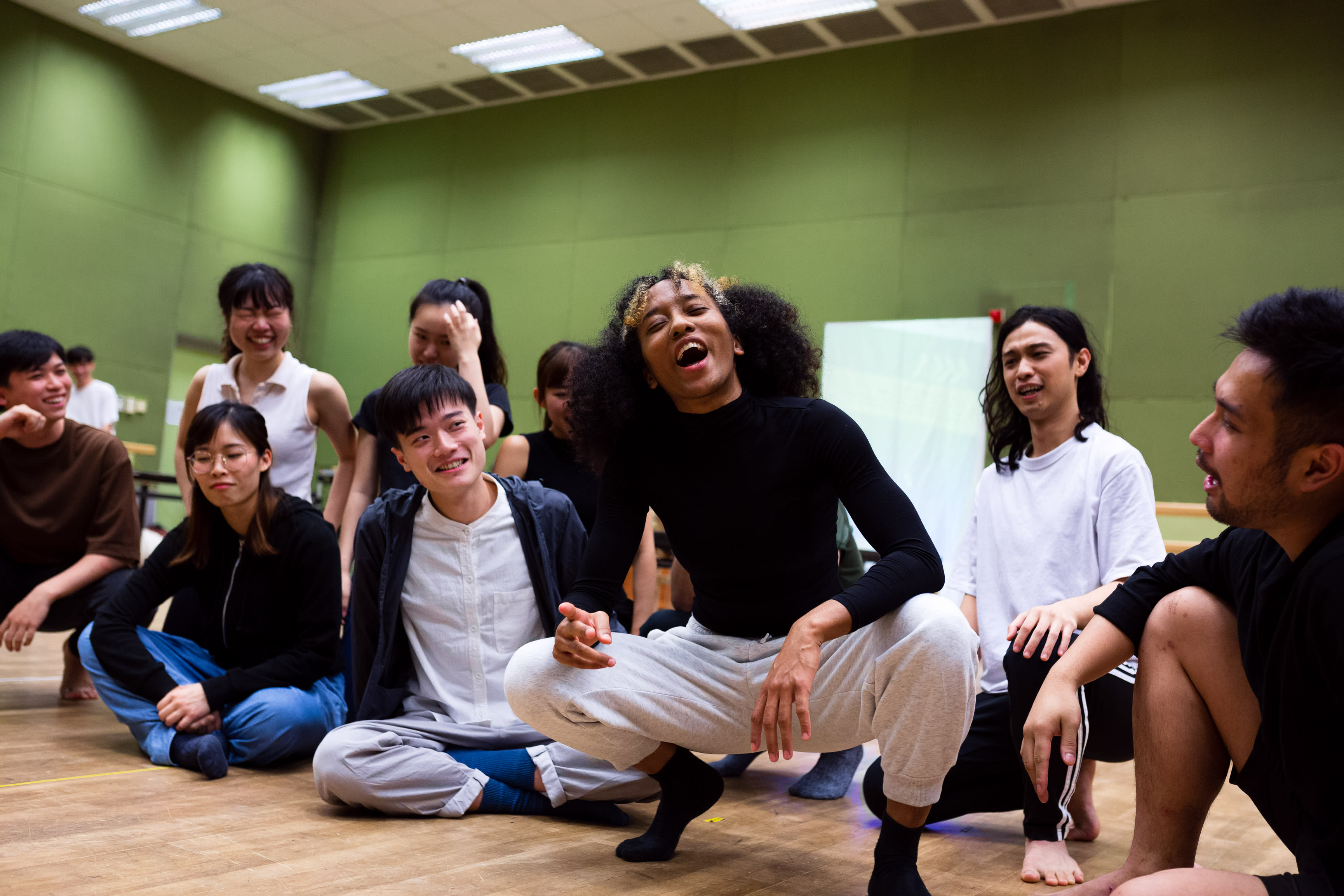 In a rehearsal for Rooftop Productions' contemporary theatre piece 'Songs of Innocence and Experience', a diverse cast of Hong Kong performers are gathered in a bright studio. In the center, Caroline Chan (Satan) crouches low, energetically mid-performance with a passionate expression, surrounded by ensemble members including Sin Lok-yan, Mok Kok-pong, and Mak Ho-tin, who look on with laughter and engagement. This rehearsal session highlights the collaborative spirit of experimental HK theatre. 'Songs of Innocence and Experience Rehearsal'