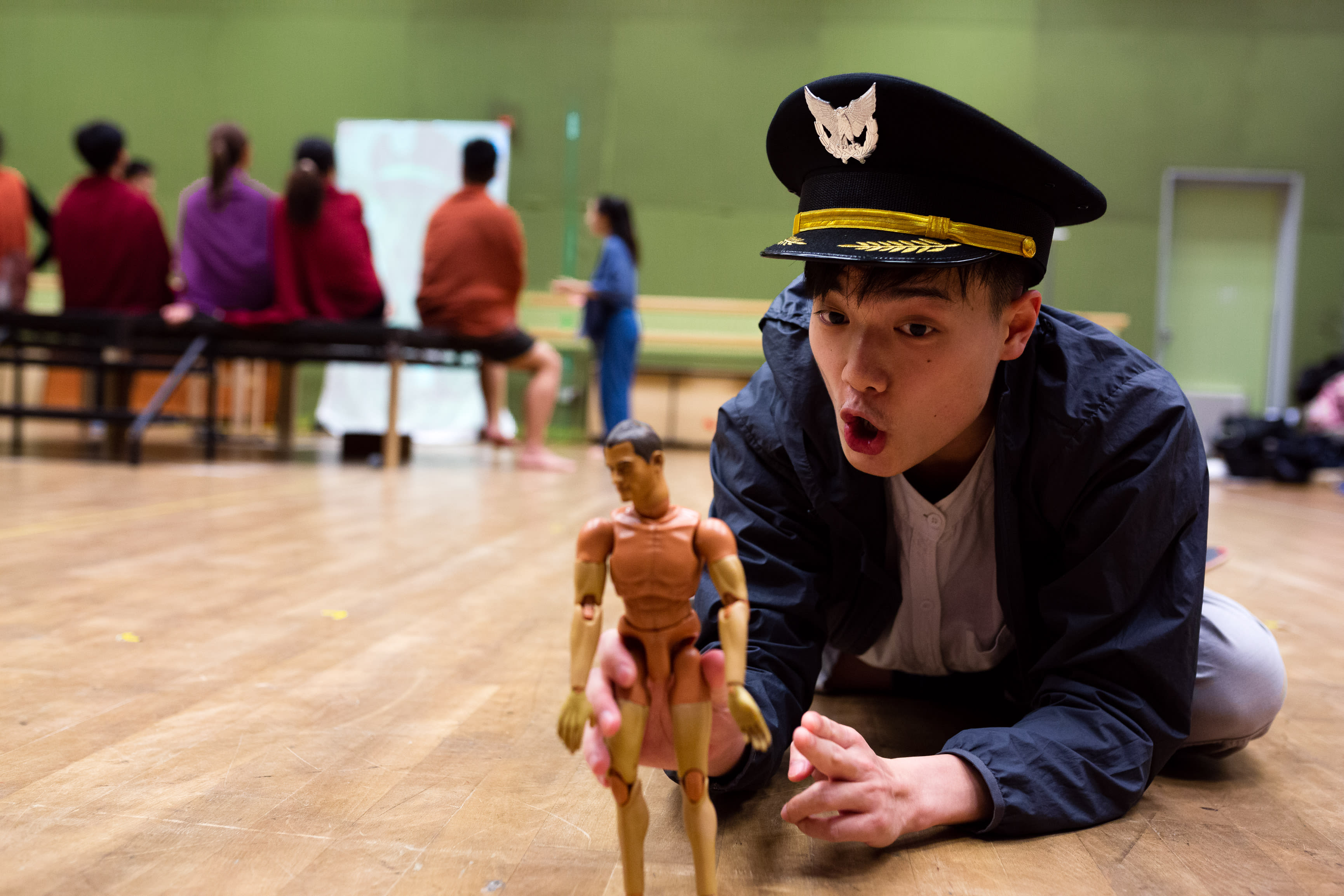 In a rehearsal for Rooftop Productions' contemporary theatre piece 'Songs of Innocence and Experience', performer Lo Man-chak (playing himself and God) kneels on a wooden stage floor. He wears a dark jacket and a formal peaked cap with a military-style emblem, looking down at a small, faceless, tan-colored articulated mannequin with an expression of intense, wide-eyed fascination. In the softly lit background, other actors in casual attire sit on a bench in front of a white projection screen, observing the creative process. Songs of Innocence and Experience Rehearsal.