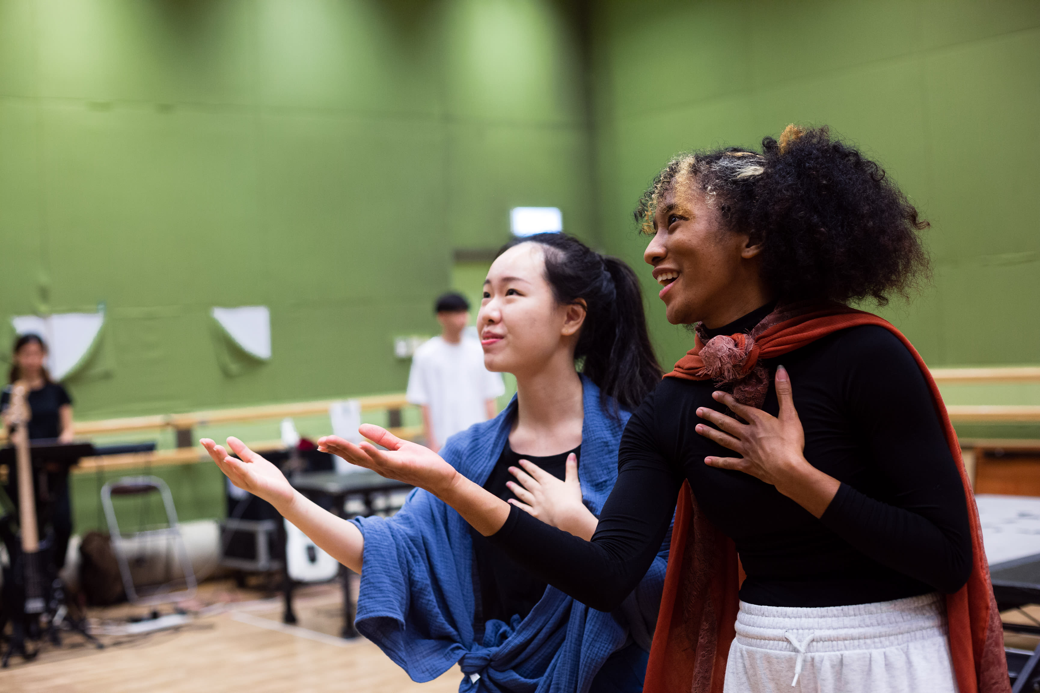 In a rehearsal for the contemporary Hong Kong theatre production 'Songs of Innocence and Experience', actors Sum Cheuk-yiu (playing Abdiel) and Caroline Chan (playing Satan/Lucifer) stand on stage. Sum wears a blue shawl and Chan a red cloak, both performers holding one hand to their chests while gesturing forward with the other, capturing a dramatic moment of emotional intensity. The backdrop is a simple green-walled studio space typical of a local rehearsal room. 'Songs of Innocence and Experience Rehearsal'