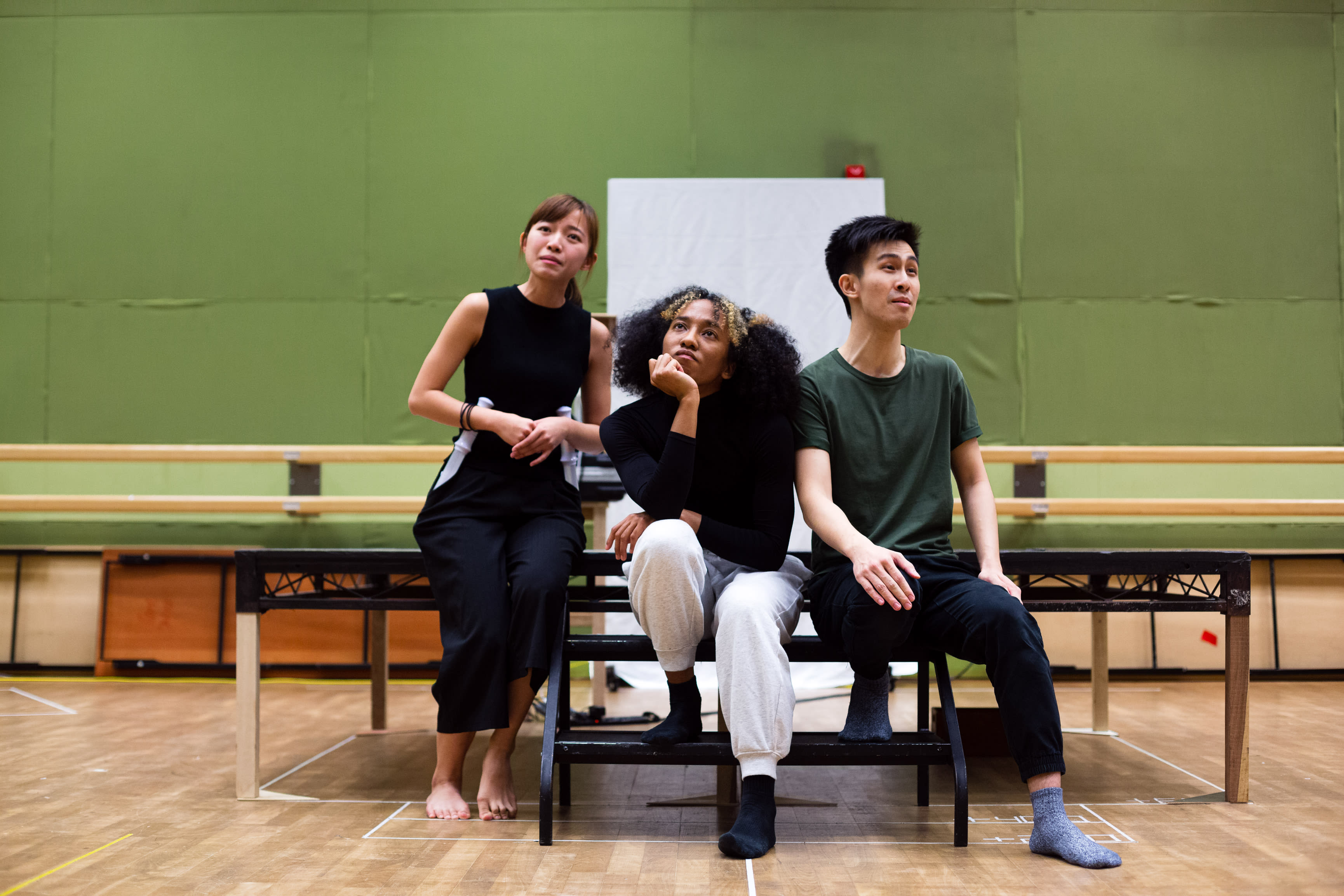 來自香港當代劇場團體「天台製作」(Rooftop Productions) 的三位演員，正為演出《天真與世故之歌》(Songs of Innocence and Experience) 進行排練。左側的冼樂欣（飾演夏娃／天使）仰頭沉思；中間的陳琬瑜（飾演撒旦／路西法）托腮思考；右側的張卓行（飾演天使）則坐姿端正，望向前方。場景設於簡約的排練室中，背後是綠色背景幕，展現了這部香港舞台製作的實驗性與排練現場的張力。《天真與世故之歌》排練