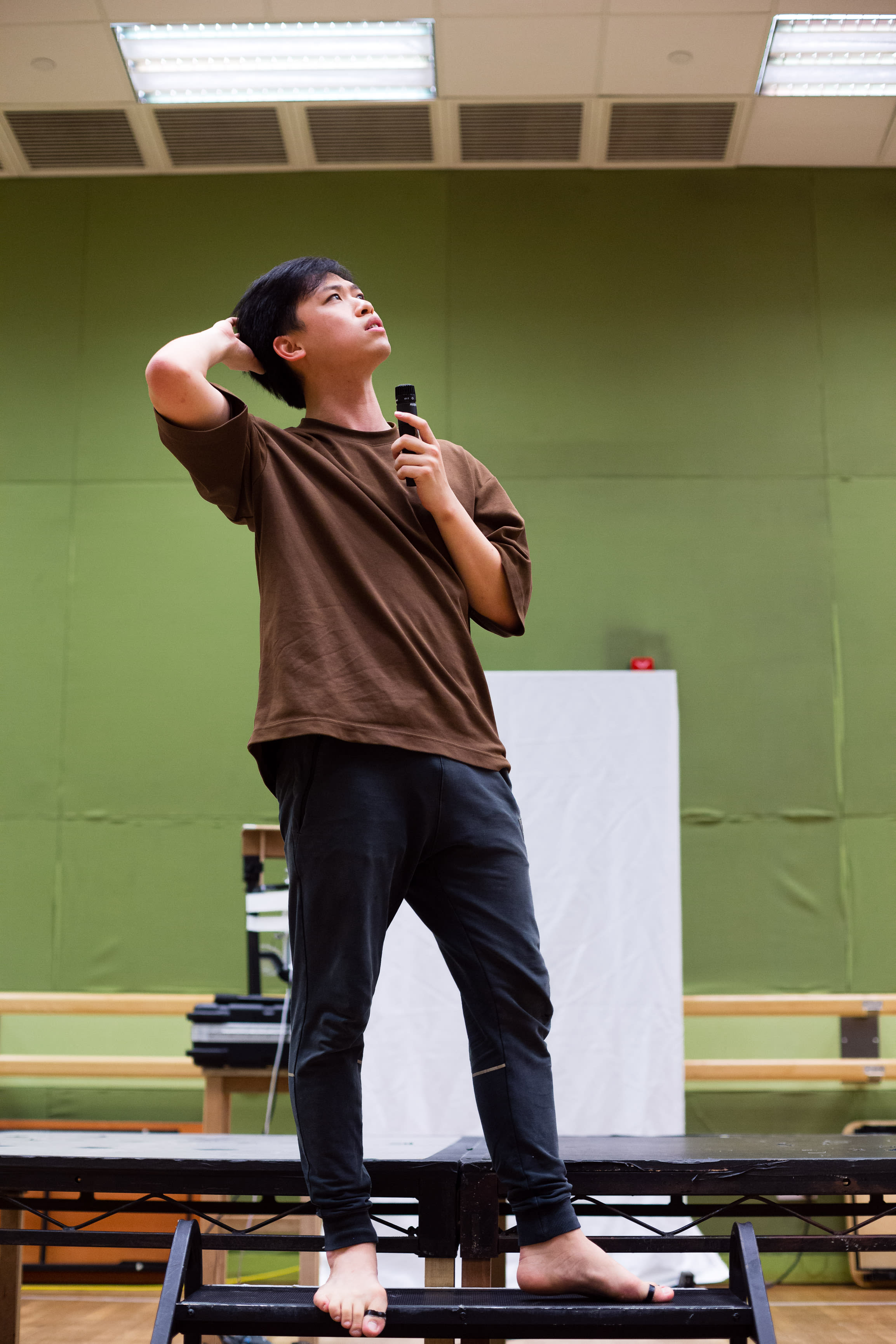 In a rehearsal for the contemporary Hong Kong stage production 'Songs of Innocence and Experience' by Rooftop Productions, actor Chew Yu-yeung (performing as Mammon) stands barefoot on a low stage platform. He is wearing a casual brown t-shirt and dark joggers, holding a microphone in one hand while his other hand rests behind his head, his gaze directed upward with an expression of contemplative inquiry. The scene is set in a studio with green acoustic wall panels and stage lighting fixtures visible above. Songs of Innocence and Experience Rehearsal.