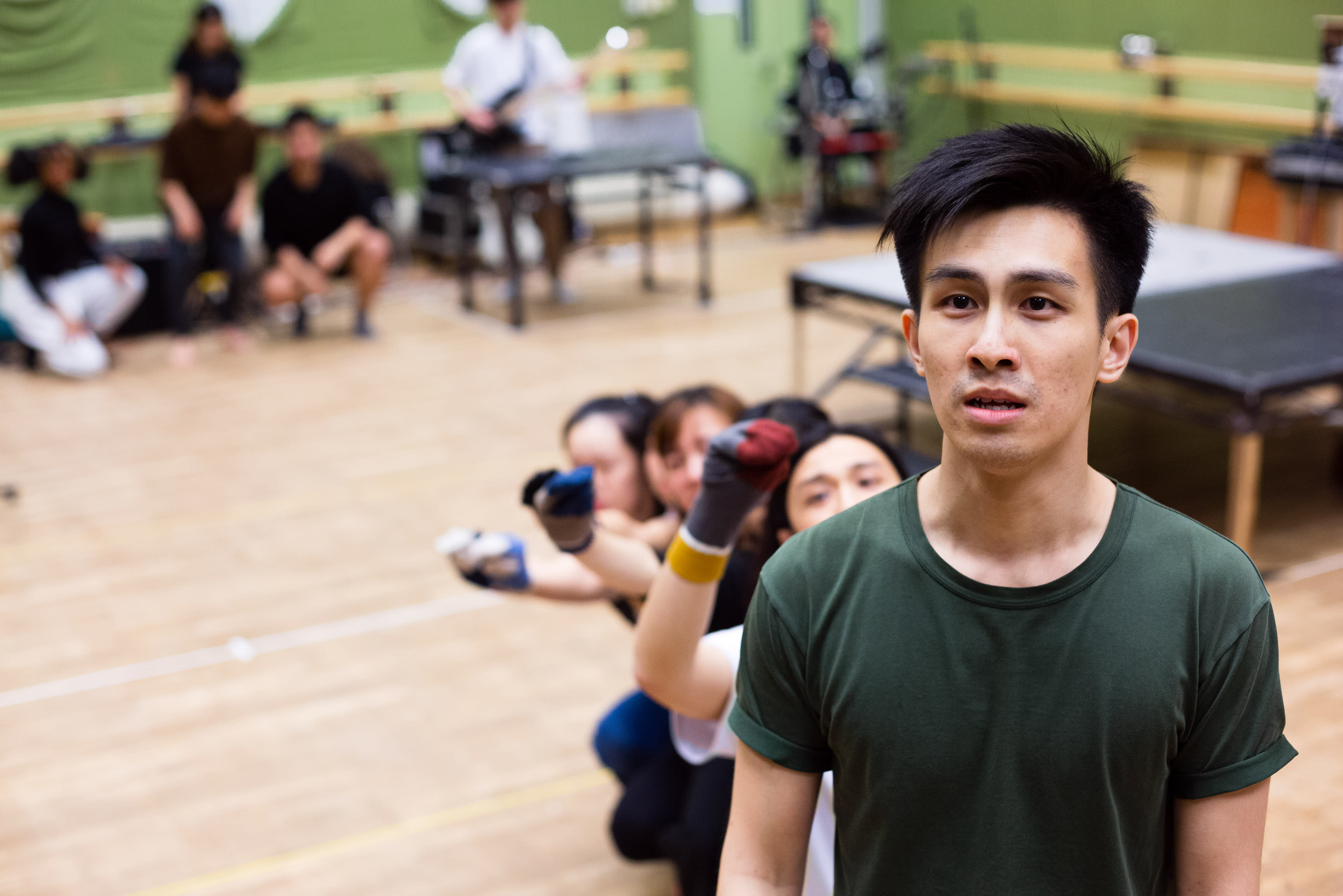 在天台製作（Rooftop Productions）當代劇場作品《天真與世故之歌》的排練現場中，演員張卓行佇立於前景，表情專注而凝重。在他身後，包括麥晧鈿在內的一眾演員排成一列，透過層層疊疊的手部動作，呈現出充滿意象的肢體編排或集體定格。排練室背景僅有簡約木凳與樂器，真實呈現香港劇場排練時那份原始且實驗性的張力。《天真與世故之歌》排練。