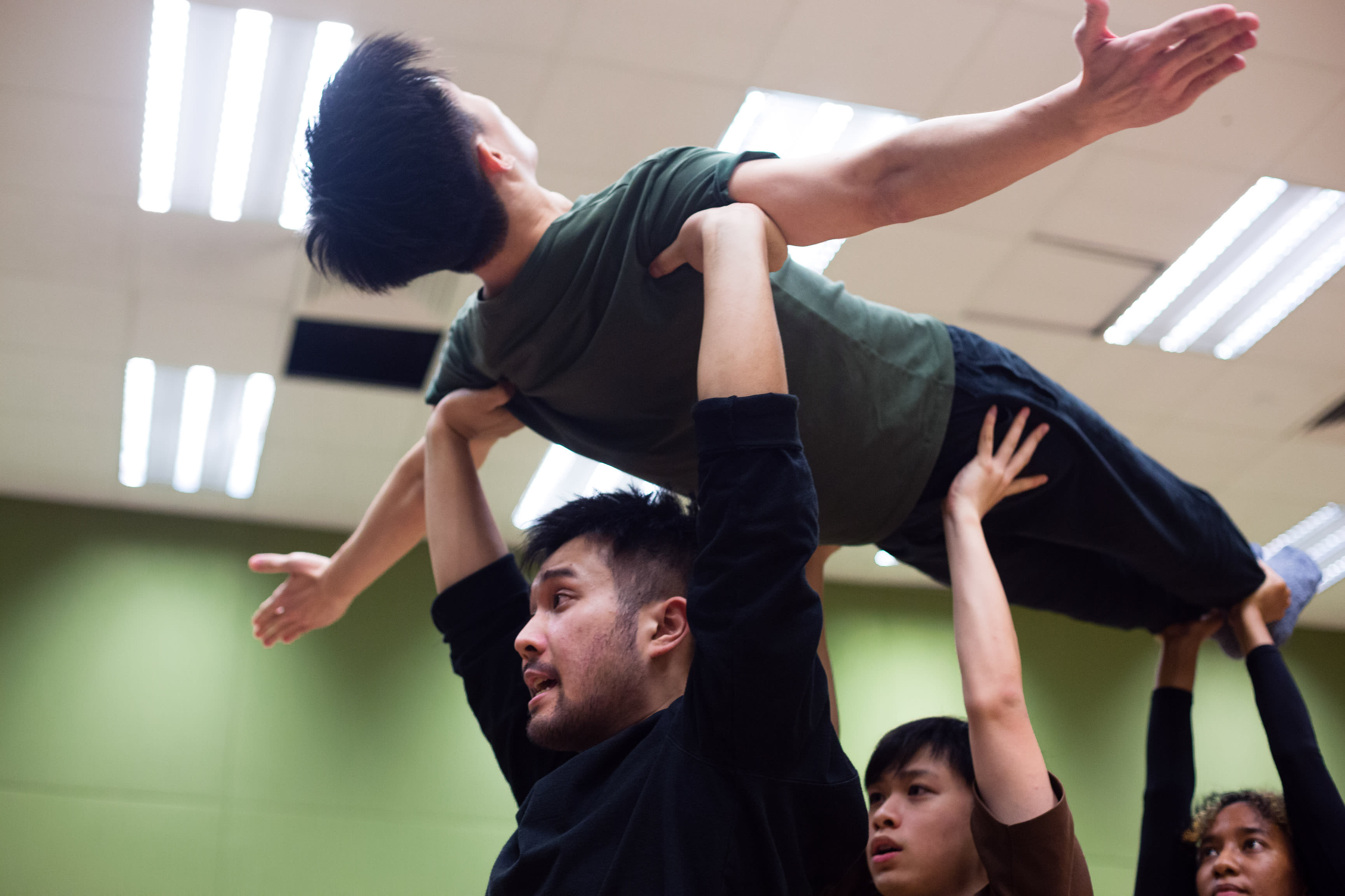 In a rehearsal for the contemporary Hong Kong theatre production 'Songs of Innocence and Experience', Mok Kok-pong (as Michael) is held aloft by fellow actors in a dynamic stage lift. Below him, Chew Yu-yeung (as Mammon) supports his torso with intense focus, while Caroline Chan (as Satan/Lucifer) assists with the lift from below. The scene is set in a studio with bright overhead panel lighting, capturing the raw energy and physicality of professional stage performance. 'Songs of Innocence and Experience Rehearsal'