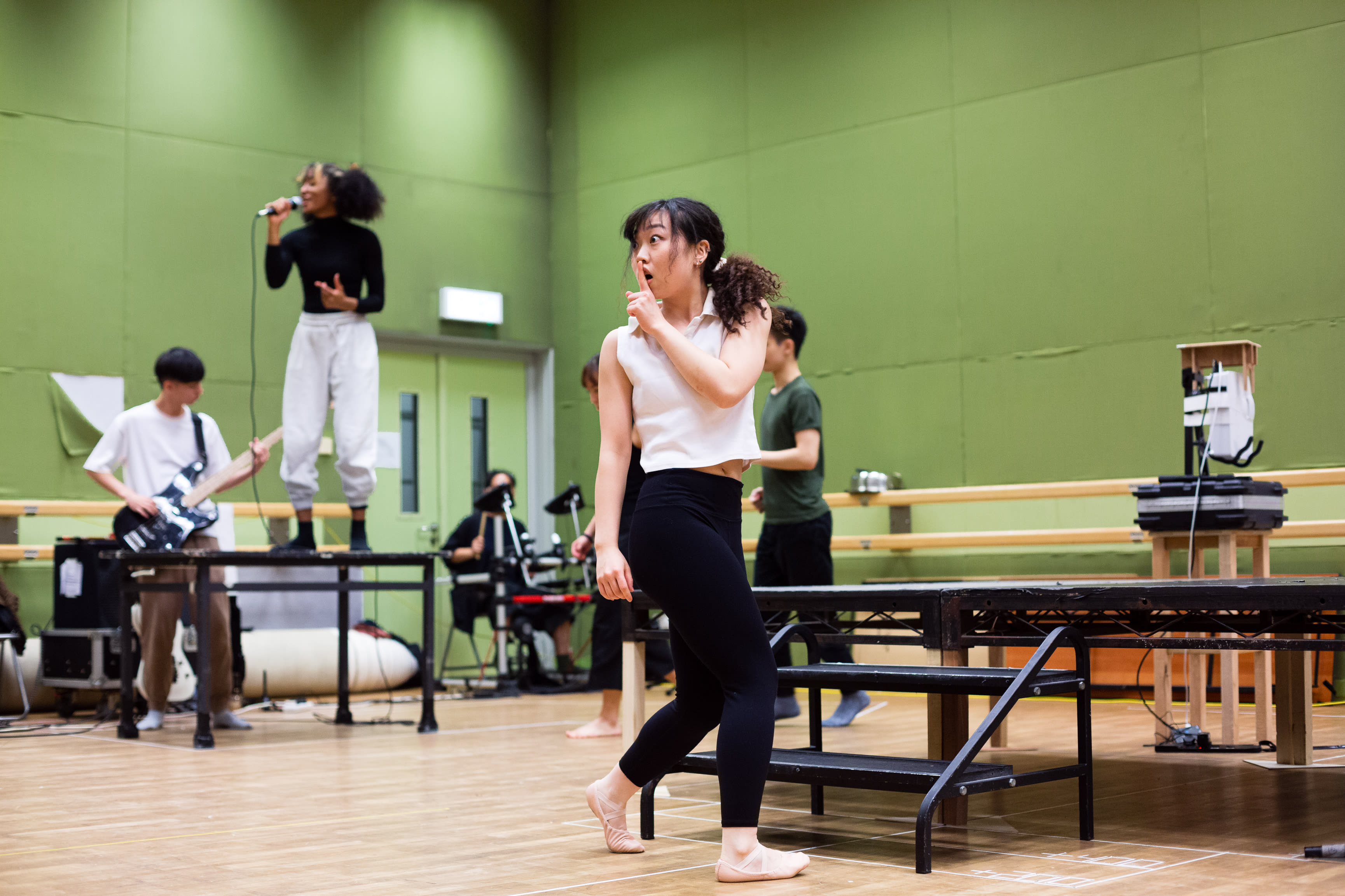 In a rehearsal for Rooftop Productions' contemporary theatre piece 'Songs of Innocence and Experience', a performer gestures with a 'shush' signal to the audience while walking across a studio stage. In the background, a vocalist performs into a microphone while standing atop a table, accompanied by a guitarist and a drummer, capturing the raw, experimental spirit of Hong Kong stage performance. Songs of Innocence and Experience Rehearsal