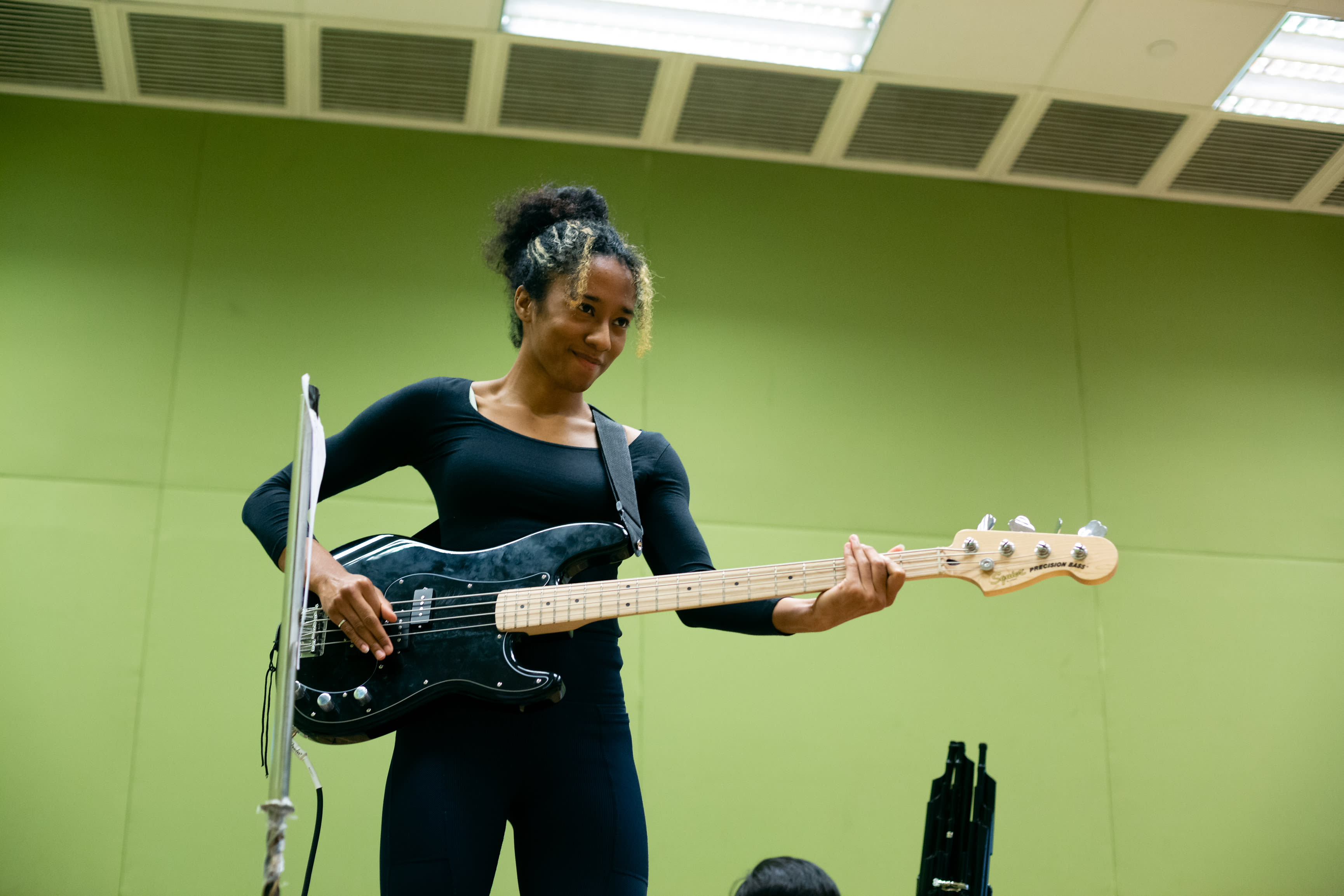 Caroline Chan, cast as Satan (Lucifer) in Rooftop Productions' contemporary theatre piece 'Songs of Innocence and Experience', plays a black electric bass guitar during a rehearsal. She is centered in the frame, wearing a black top and looking down at the instrument with a focused, gentle expression against a plain light green backdrop. This intimate rehearsal capture highlights the artistic process behind this Hong Kong stage performance. Songs of Innocence and Experience Rehearsal.