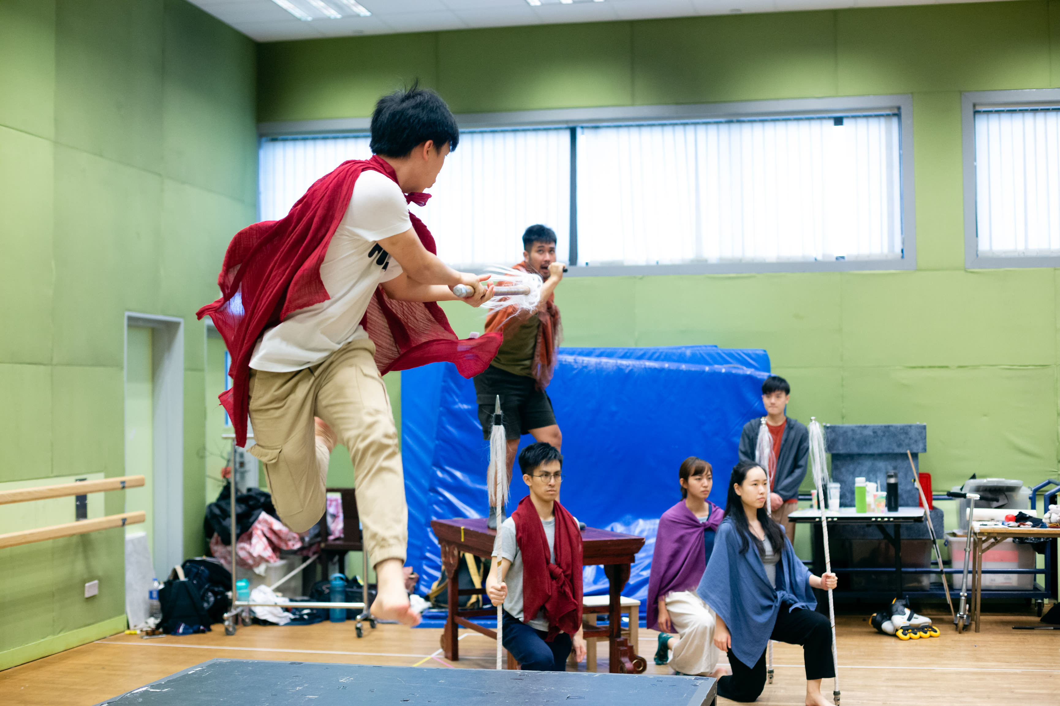 In a dynamic rehearsal for Rooftop Productions' contemporary theatre piece 'Songs of Innocence and Experience' (《天真與世故之歌》), an actor playing an Angel performs a dramatic, mid-air leap across the stage, wearing a flowing red cape. Behind him, another performer playing God stands atop a wooden platform, while cast members, including those portraying Michael, Mammon, Eve, and Abdiel, sit nearby in a bright, modern studio space. The rehearsal captures the physical intensity and raw artistry characteristic of Hong Kong's independent theatre scene. 'Songs of Innocence and Experience Rehearsal'