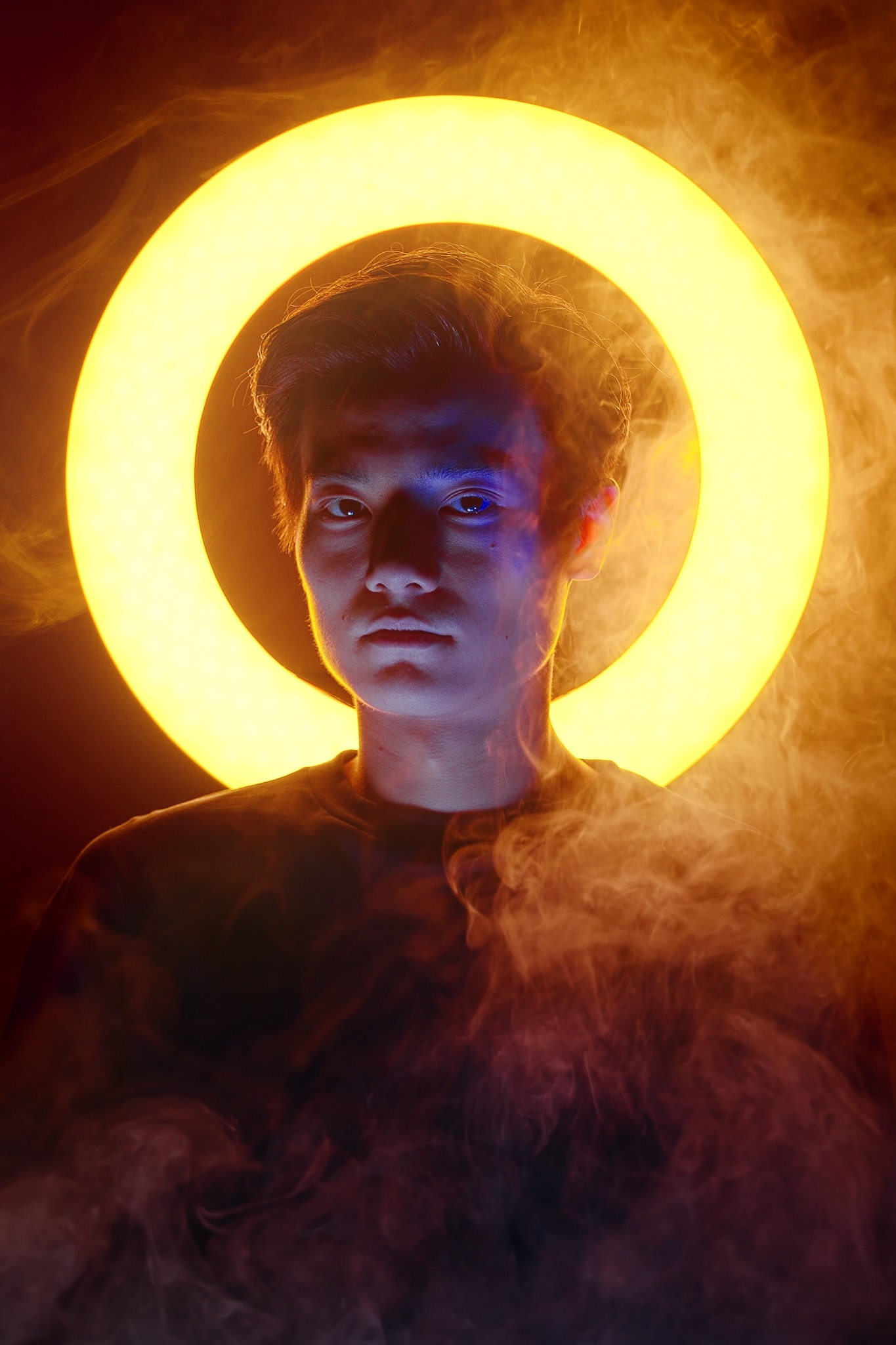 A dramatic promotional portrait for the contemporary Hong Kong theatre production 'Songs of Innocence and Experience' (WestK) by Rooftop Productions, featuring actor Lo Man-chak. He is centered in the frame, cast in a striking dual-lighting setup: a bright, circular halo of golden light behind his head, contrasting with a cool blue spotlight on his face, creating an ethereal, god-like silhouette. Thick, swirling smoke envelops the lower half of his body, adding a sense of mystery to this HK stage performance. 'Lo Man-chak, Promo for Songs of Innocence and Experience (WestK)'