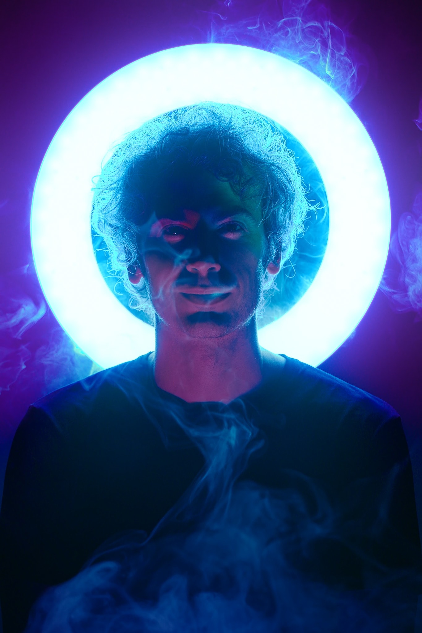 A dramatic headshot of actor Ivor Houlker for the contemporary Hong Kong theatre production 'Songs of Innocence and Experience' at WestK. He is centered against a vibrant, glowing circular blue neon light that frames his head like a halo, with swirling, atmospheric smoke foregrounding his dark shirt, evoking a mysterious and introspective mood characteristic of modern stage performance. Ivor Houlker, Promo for Songs of Innocence and Experience (WestK).