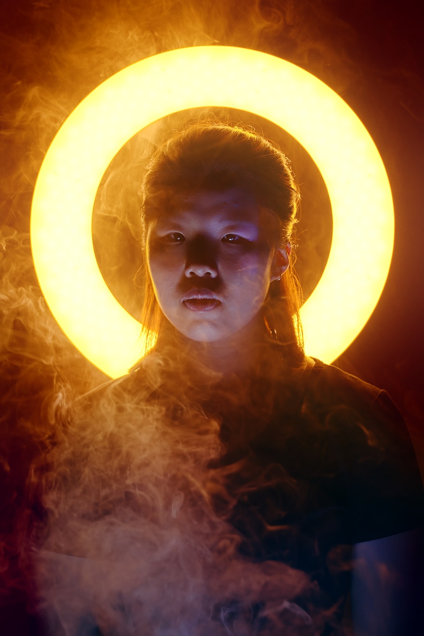 顏詩鏝（Tiffany）在天台製作的當代劇場作品《天真與世故之歌》中，置身於繚繞的煙霧中，呈現出引人深思的舞台神態。表演者身後是一道奪目的環形背光，營造出空靈的氛圍，突顯出戲劇張力。強烈的燈光與煙霧效果，展現了這部西九文化區（WestK）演出作品的前衛實驗風格。『顏詩鏝，天真與世故之歌（西九文化區）』