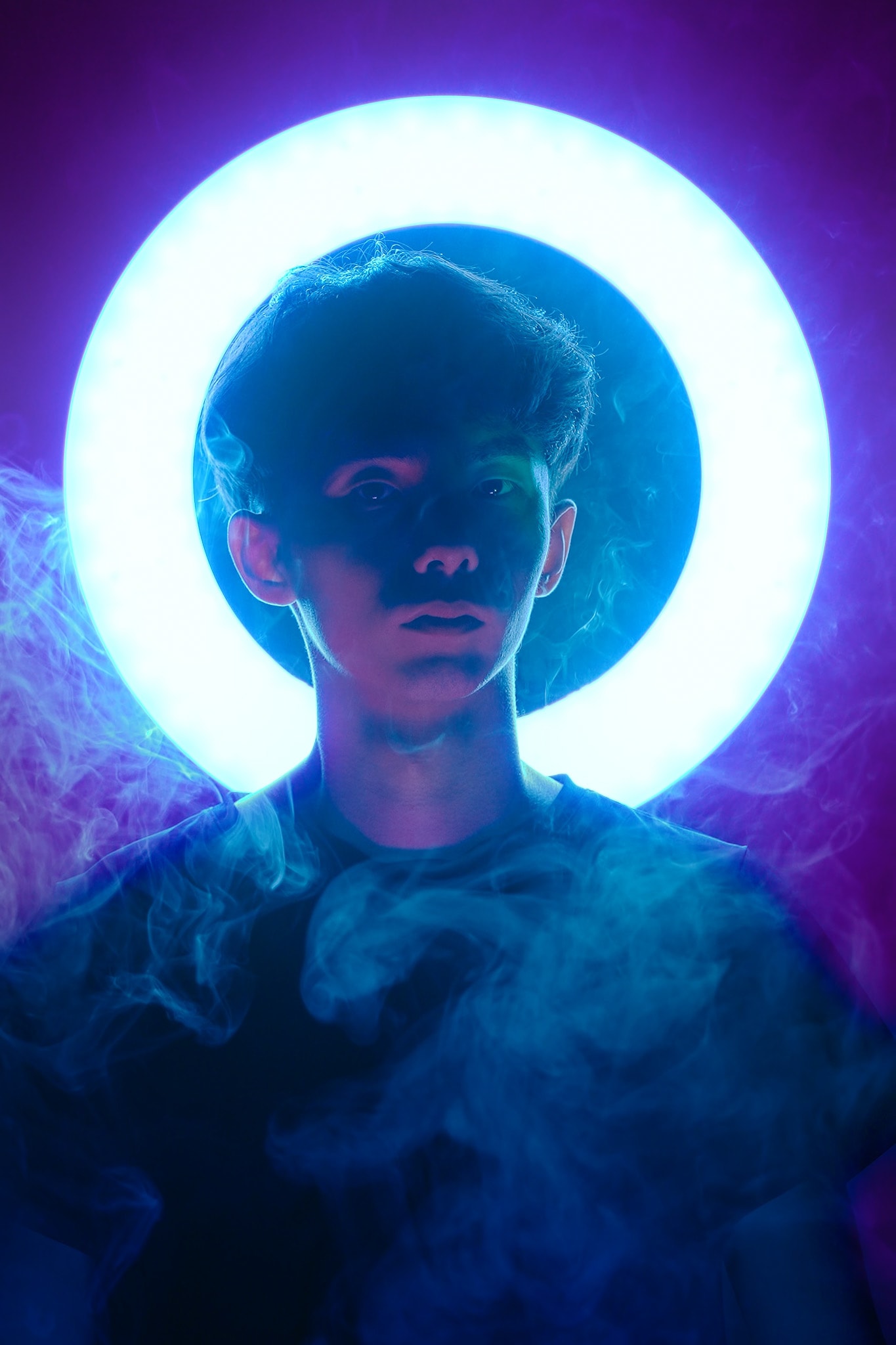 A dramatic promotional portrait of actor Wu Lui-fung for the contemporary Hong Kong theatre production 'Songs of Innocence and Experience' at WestK. The image features a medium close-up of Wu, lit by a vibrant, circular neon-blue ring light positioned directly behind his head, creating a halo effect. He stands amidst wispy, atmospheric stage smoke that obscures the lower half of his torso, cast in moody blue and magenta hues. Wu's expression is intense and contemplative, embodying the experimental aesthetic of Rooftop Productions (天台製作). Wu Lui-fung, Promo for Songs of Innocence and Experience (WestK)