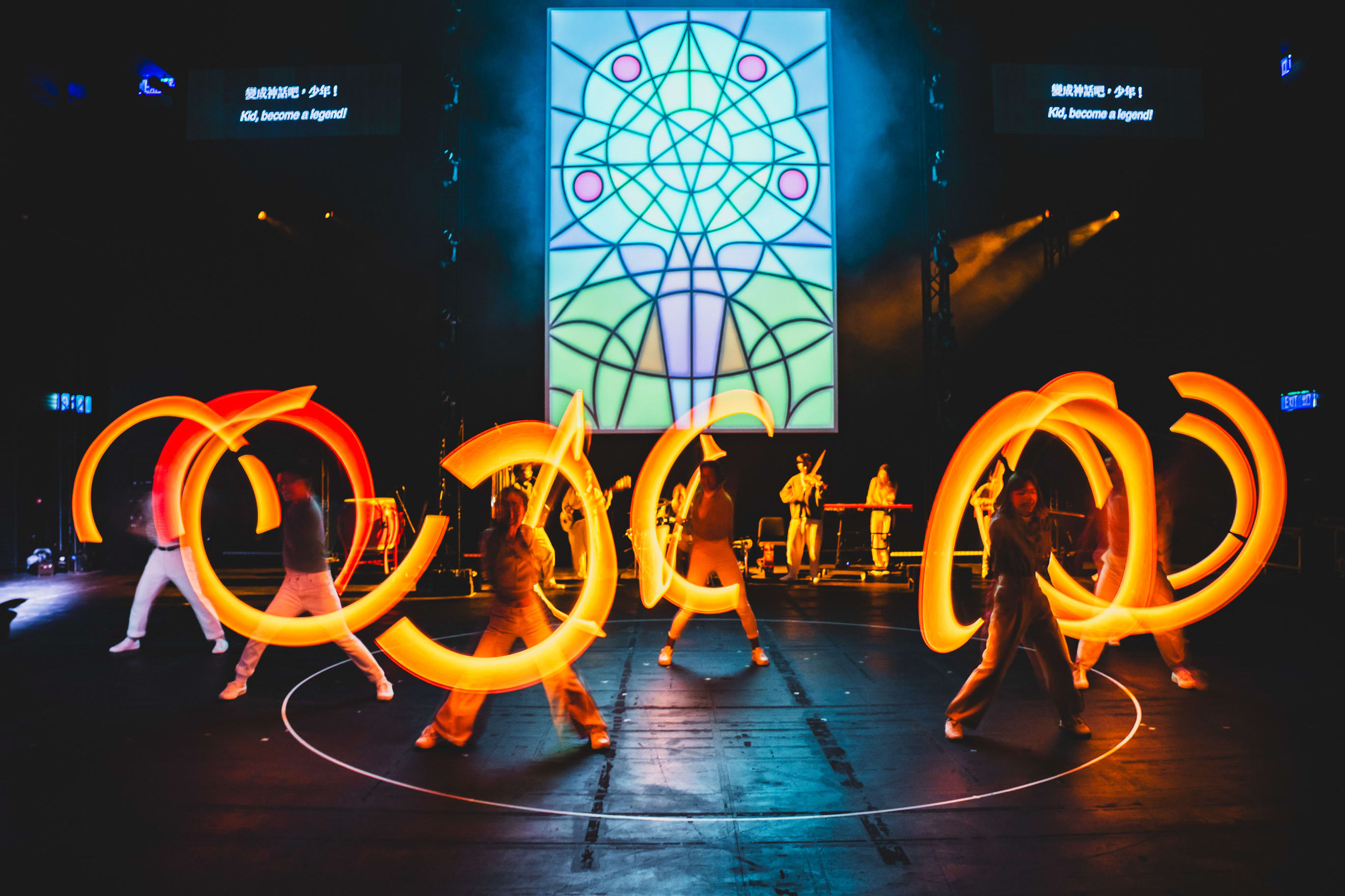 Performers on stage at WestK for Rooftop Productions' contemporary theatre piece, Songs of Innocence and Experience, moving in a circle while swinging vibrant, glowing orange light tubes that create long-exposure light trails. The dramatic lighting highlights the performers' dynamic movements against a dark stage, underscored by a large, geometric, neon-lit stained glass backdrop. This visually arresting scene captures the energy of Hong Kong's independent theatre scene. 'Songs of Innocence and Experience (WestK).'