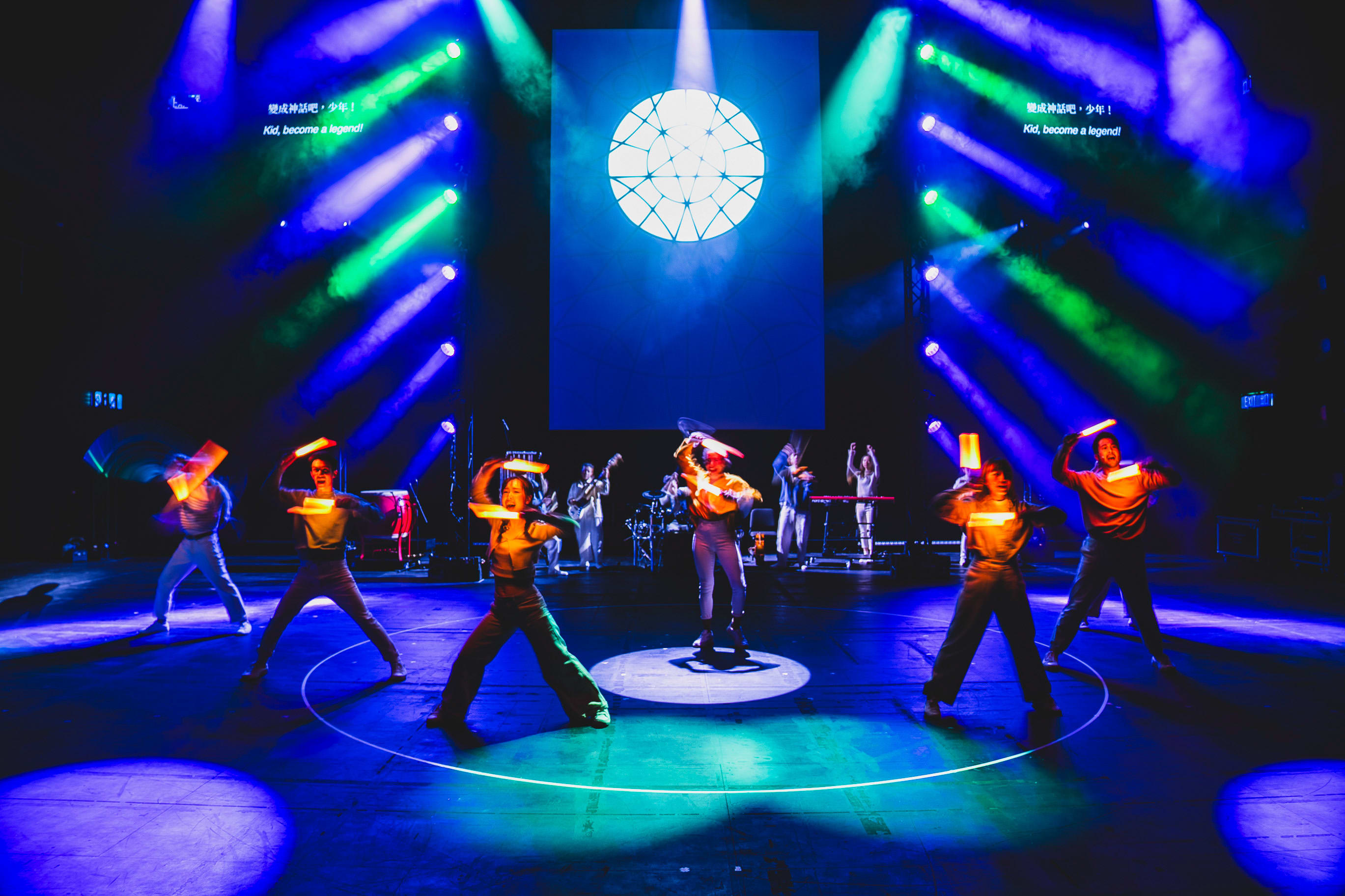 A vibrant wide shot of a contemporary stage performance for 'Songs of Innocence and Experience' by Rooftop Productions at WestK. In the center, a large, glowing circular light projection mimics a sun or celestial sphere against a deep blue backdrop. Six performers are positioned on stage in dynamic poses, holding glowing orange LED light bars that cut through the atmospheric haze and blue stage lighting. The performers, including Caroline Chan, Mok Kok-pong, Sin Lok-yan, Cheung Cheuk-hang, and Sum Cheuk-yiu, wear modern attire as they embody their roles. The scene captures a powerful moment of movement and light in this Hong Kong experimental theatre production. 'Songs of Innocence and Experience (WestK)'