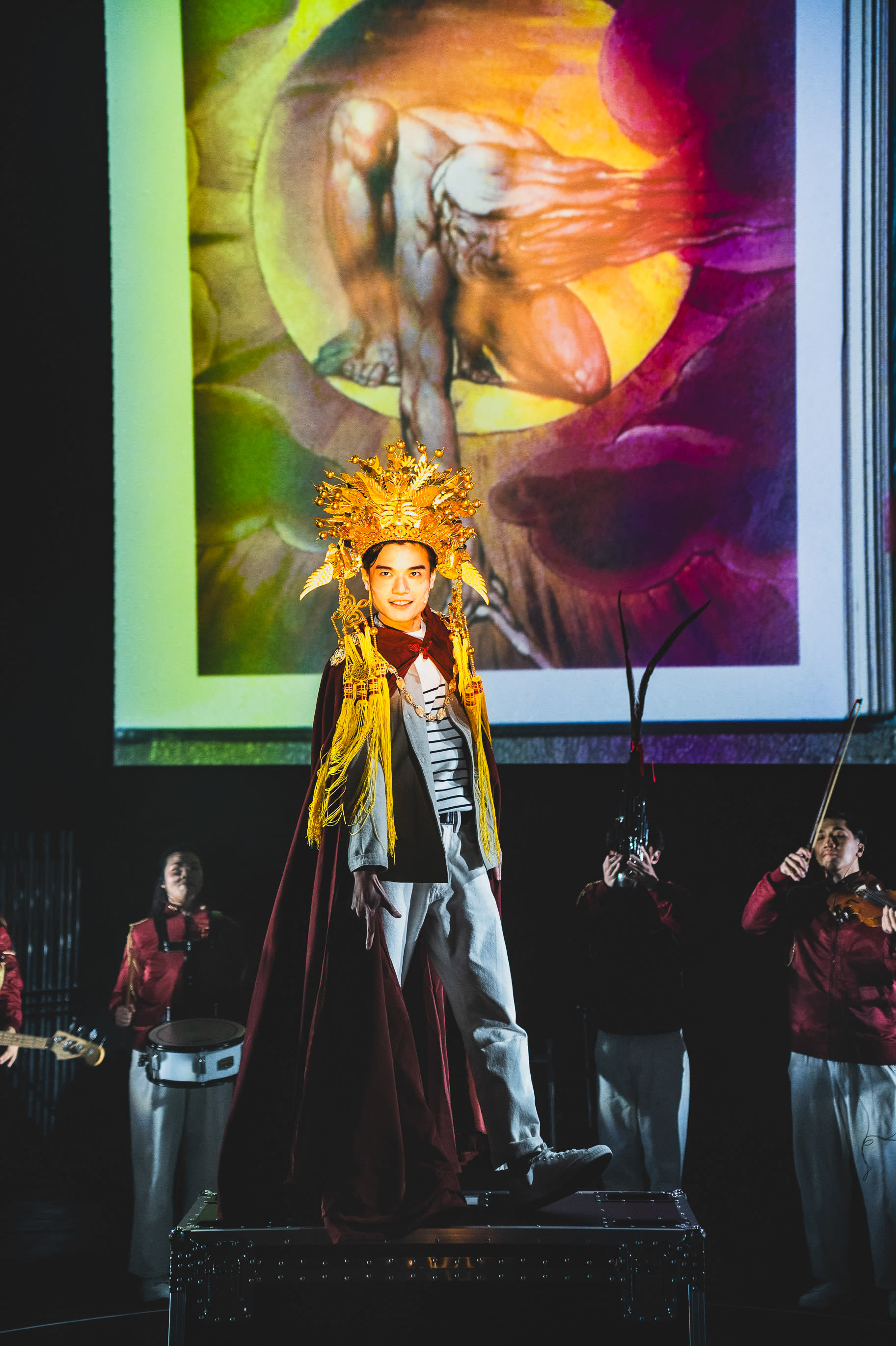 A dramatic stage shot from the contemporary Hong Kong theatre production 'Songs of Innocence and Experience' by Rooftop Productions (天台製作). At the center, Lo Man-chak, portraying 'God,' stands confidently on a black road case, wearing a majestic, intricate gold crown and a deep maroon cape over contemporary clothing. Behind him, a large projection features a surreal, muscular figure curling within a golden sphere. To his sides, Ngan Sze Man Tiffany and Kwok Chun Hin Justin, dressed in matching red jackets and white trousers, provide musical accompaniment on instruments including a snare drum and violin. The lighting is moody and theatrical, highlighting the contrast between the celestial backdrop and the performers on stage. 'Songs of Innocence and Experience (WestK)'