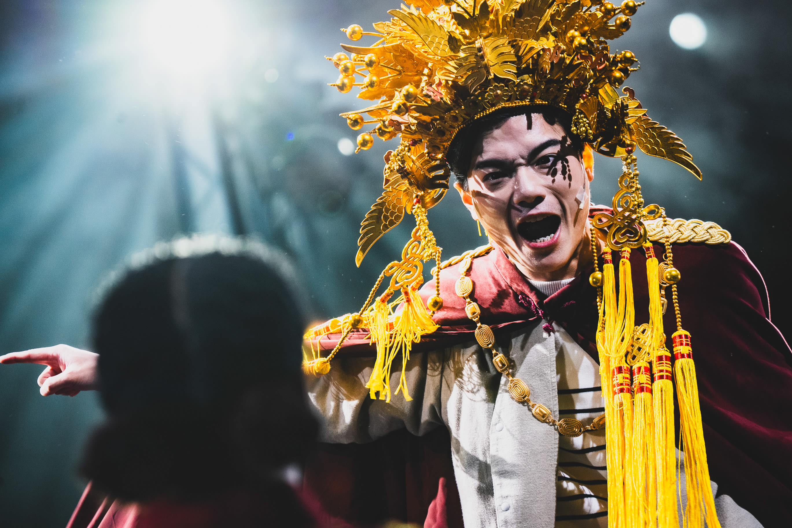 天台製作（Rooftop Productions）當代劇場作品《天真與世故之歌》於西九文化區的演出劇照。演員羅文澤飾演「上帝」，頭戴誇張金飾頭冠，身披深紅色斗篷，神情激昂地指向舞台另一側，展現出強烈的戲劇張力。舞台燈光在角色背後營造出聚光效果，突顯這部香港舞台劇的演出氛圍。《天真與世故之歌》（西九）