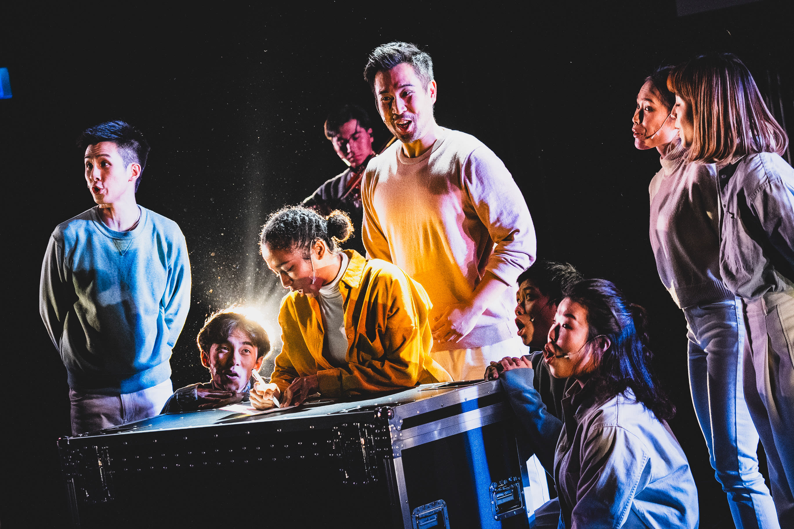 A dramatic stage scene from the Rooftop Productions (天台製作) performance, 'Songs of Innocence and Experience' (西九). Under moody, high-contrast spotlighting, an ensemble of performers gathers around a large, black industrial equipment case. A young woman in a vibrant yellow jacket leans over the case, writing intensely as a beam of light illuminates the area, while fellow performers—including Mok Kok-pong, Caroline Chan, and others—watch with expressions ranging from concern to anticipation. This contemporary Hong Kong theatre production blends experimental performance with evocative staging. 'Songs of Innocence and Experience' (WestK)