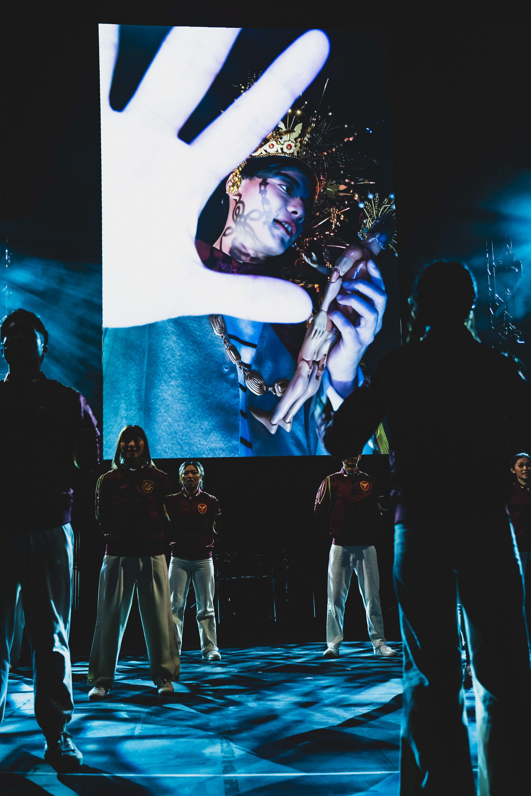 A dramatic stage scene from the Rooftop Productions contemporary theatre work 'Songs of Innocence and Experience' at WestK. In the foreground, a group of performers stand in semi-silhouette, illuminated by cool, blue stage lights that cast intricate patterns on the floor. Behind them, a large LED screen displays a striking close-up of actor Chin Wing-kar in the role of Belial, wearing an ornate, spiky headpiece and dramatic makeup, with a giant, glowing hand gesture reaching toward the foreground, creating a surreal and otherworldly atmosphere. 'Songs of Innocence and Experience (WestK)'