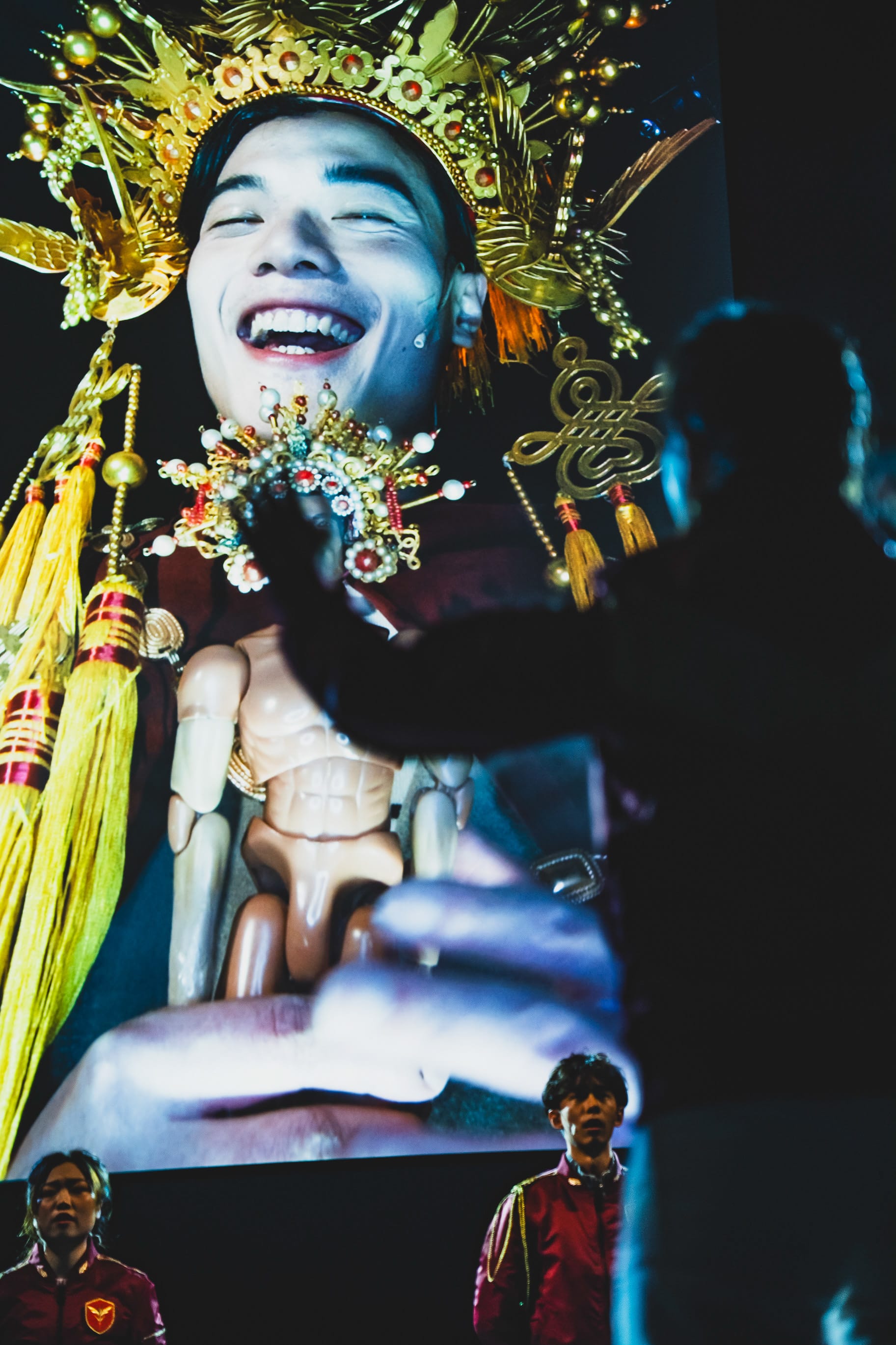 天台製作（Rooftop Productions）於西九文化區演出的《天真與世故之歌》舞台劇照。舞台背景是一幅巨大的高對比投影，畫面中的羅文澤佩戴著華麗的金色傳統頭飾與飾品，面帶燦爛笑容，手中捧著一尊關節可動的人偶。舞台前景可見一名表演者的剪影向投影伸出手，而在下方，兩位身穿深紅色制服的表演者——鄔磊鋒與李安生——站在戲劇性的燈光中。此作品巧妙融合了當代香港劇場美學與儀式感的民俗視覺元素。《天真與世故之歌》（西九）