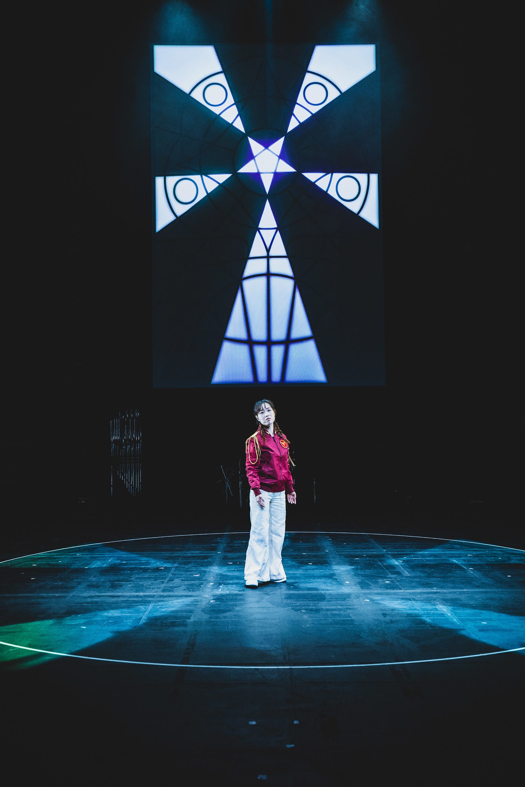 表演者沈卓瑤站在天台製作（Rooftop Productions）當代劇場作品《天真與世故之歌》（於西九文化區演出）的舞台中央。她身穿紅色飛行外套與白色長褲，置身於圓形舞台上的冷調藍色光束中。背景投影著幾何星形與羽翼般的圖案，展現出香港當代劇場獨特的先鋒美學與舞台調度。 《天真與世故之歌》（西九）