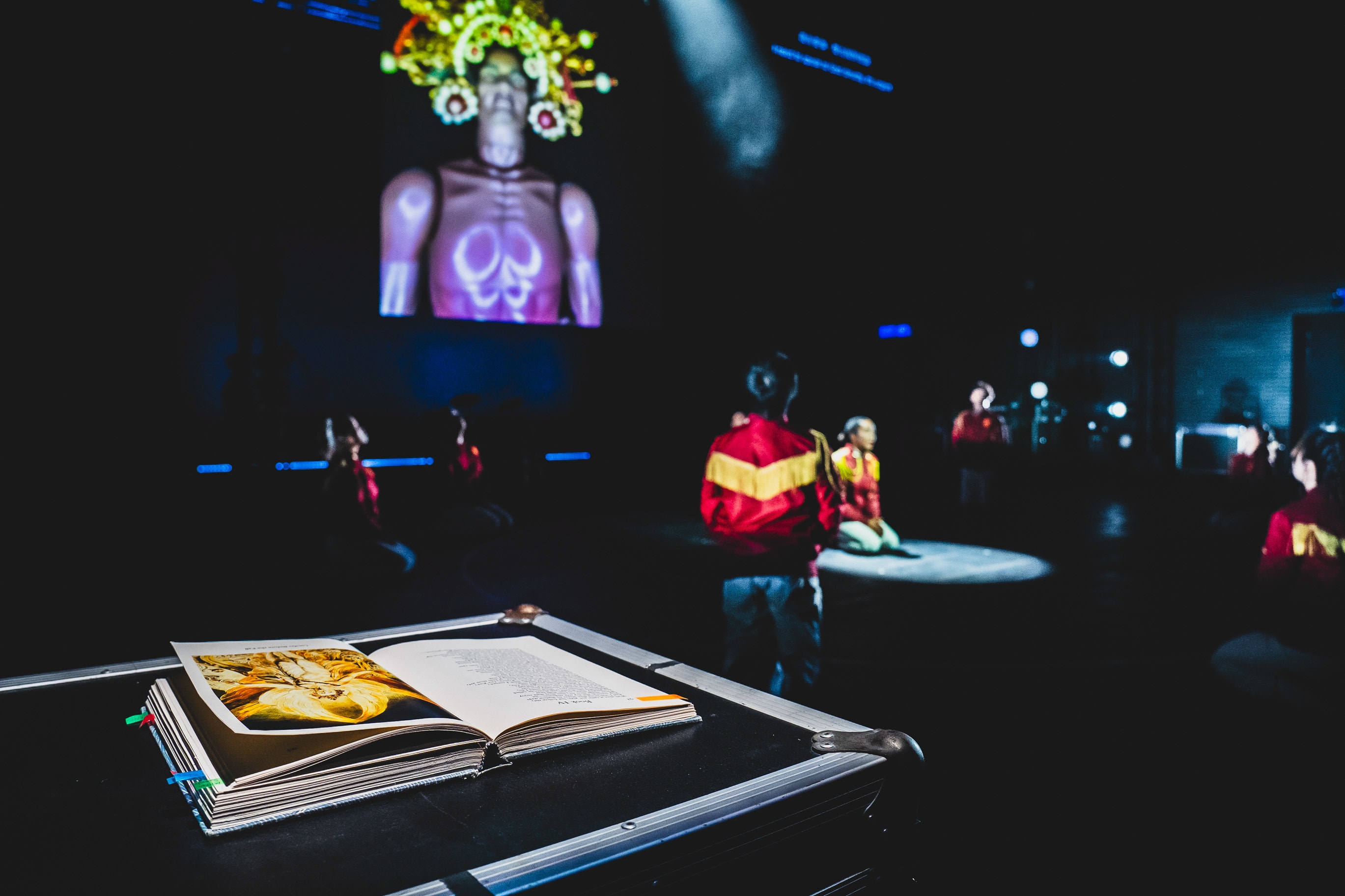 In a dark, atmospheric theatre space, a performer stands in the foreground wearing a red jacket with yellow chevrons, facing a large projection on the back wall of a figure adorned with a golden, glowing crown. In the foreground, an open book rests on a road case, its pages illuminated, suggesting an intimate connection between text and performance. This rehearsal shot of 'Songs of Innocence and Experience' highlights the experimental stage design and multimedia storytelling characteristic of Rooftop Productions' contemporary Hong Kong theatre work. 'Songs of Innocence and Experience (WestK)'