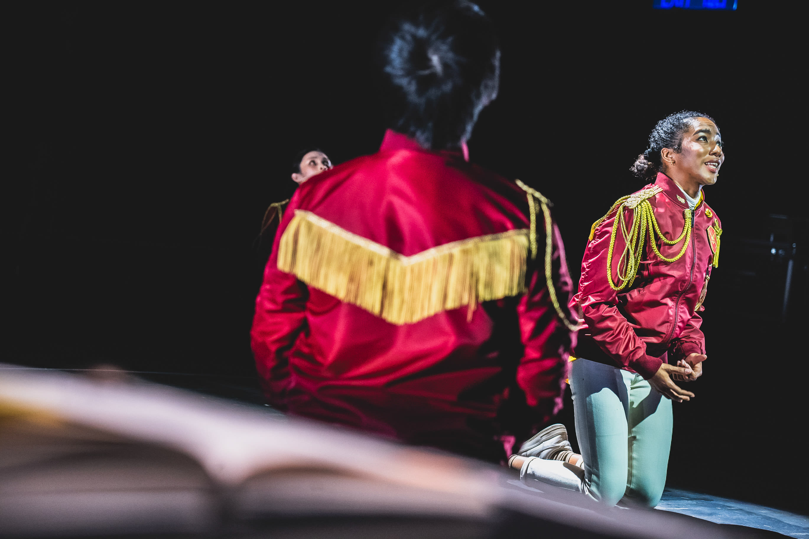 在天台製作於西九文化區上演的當代劇場作品《天真與世故之歌》中，飾演路西法的演員陳琬瑜在強烈燈光下跪於舞台。她身穿飾有金色流蘇肩章的紅色軍裝風格夾克，神情專注地向上仰望，充滿感染力。前景中，飾演彼列的演員錢穎嘉以模糊的背影入鏡，營造出豐富的舞台空間感與戲劇張力。《天真與世故之歌》（西九）