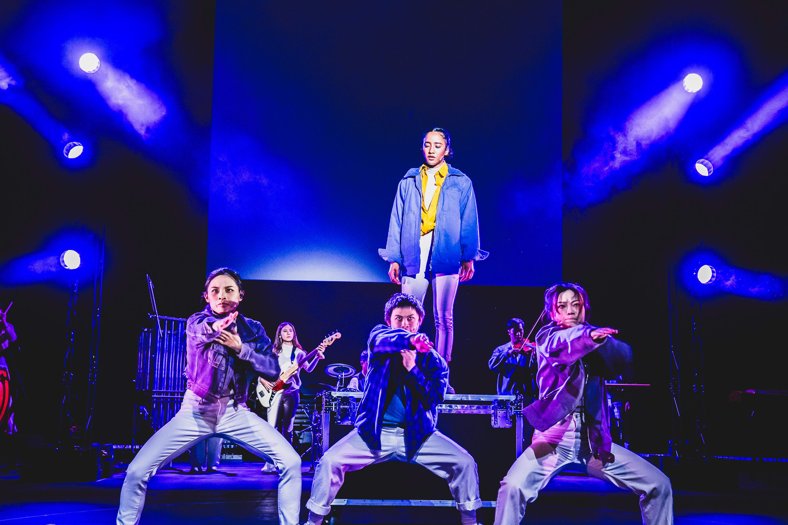 A dramatic stage shot from the contemporary theatre production 'Songs of Innocence and Experience' by Rooftop Productions at WestK. In the center, Caroline Chan stands elevated in a yellow shirt and blue jacket, looking stoic, while three performers (including Lee On-sang and Chin Wing-kar) in the foreground execute a sharp, synchronized dance pose in denim jackets and white trousers. The stage is bathed in deep, moody blue spotlights, emphasizing the high-energy, experimental atmosphere of this Hong Kong stage performance. 'Songs of Innocence and Experience (WestK)'