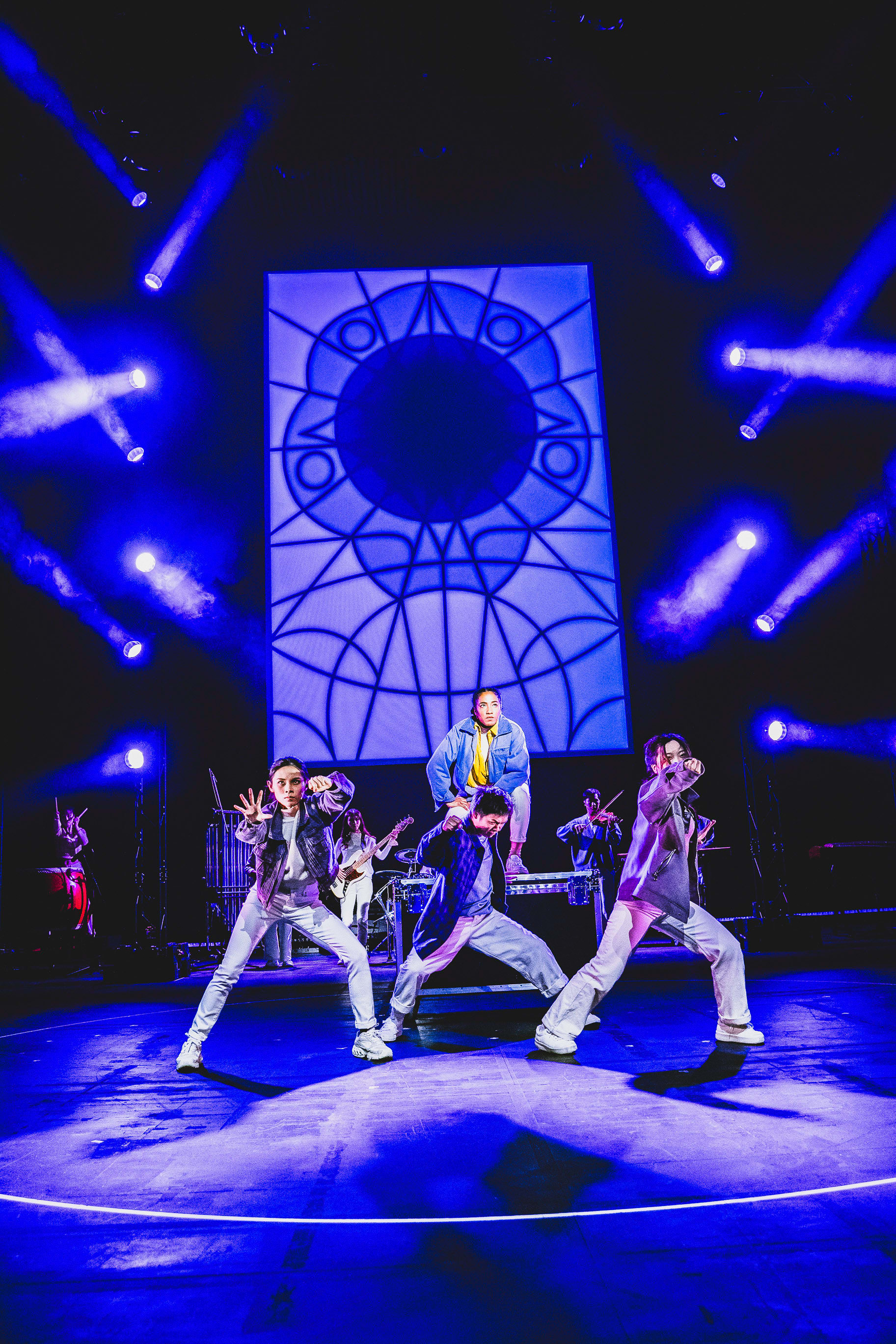 A wide-angle, low-angle shot of a dynamic contemporary dance and music performance on stage during Rooftop Productions' (天台製作) theatre piece, 'Songs of Innocence and Experience'. Three performers in the foreground are captured mid-movement: a woman in a dark jacket on the left, a man in a navy shirt crouching in the center, and a woman in a grey coat lunging to the right. Behind them, another performer stands on a raised platform, with further musicians visible in the background. The scene is illuminated by dramatic, high-contrast blue stage lighting and sharp beams that create a striking atmosphere, centered beneath a large, circular geometric backdrop. 'Songs of Innocence and Experience (WestK)'