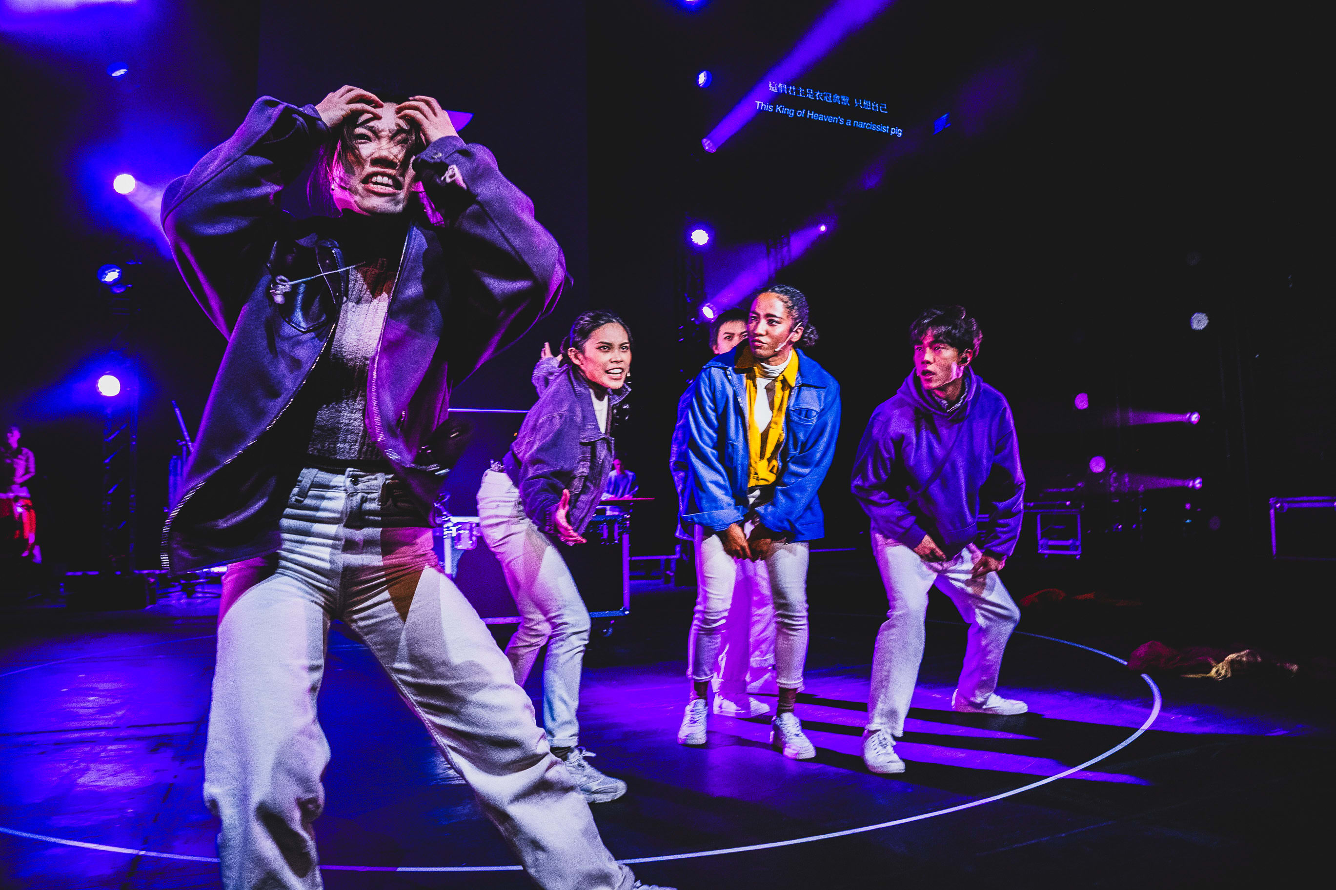 A dramatic stage shot of Rooftop Productions' contemporary theatre performance, 'Songs of Innocence and Experience', at WestK. In a wash of vibrant purple stage lighting, performer Caroline Chan (Lucifer) stands in the foreground with her hands to her head in an expression of intense anguish. Behind her, three other performers—Lee On-sang (Moloch), Chew Yu-yeung (Mammon), and Wu Lui-fung (Beelzebub)—stand in a line, captured in dynamic, focused postures during the live production. The atmosphere highlights the raw emotion and modern staging characteristic of this Hong Kong experimental production. 'Songs of Innocence and Experience (WestK)'