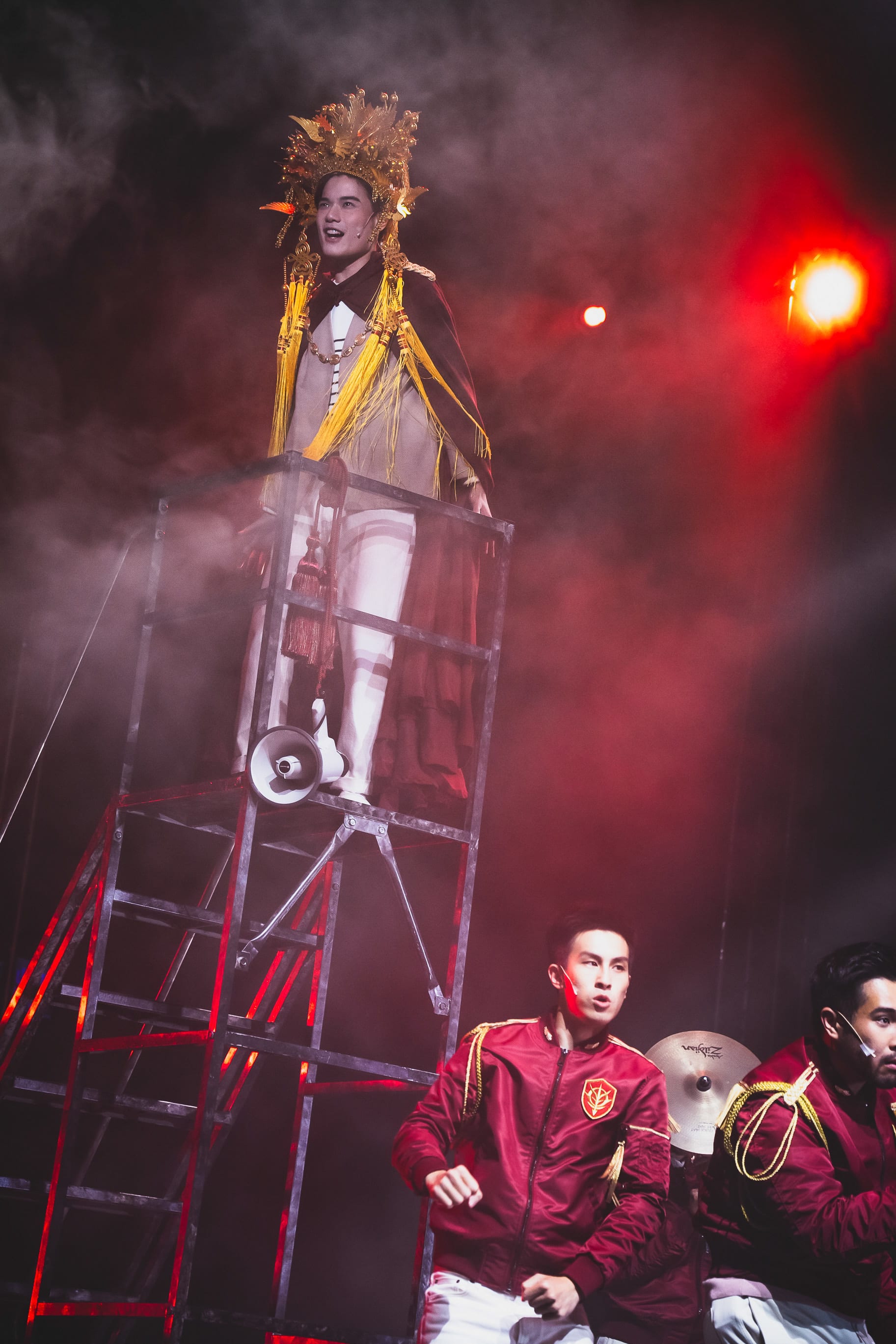 On a dramatic, dimly lit stage, a performer wearing an elaborate, shimmering gold headpiece and a mantle of gold tassels stands atop a tall, industrial metal scaffold tower, looking down and singing into a microphone. Below him, two performers in matching deep red military-style bomber jackets with gold accents and crests perform intense, focused choreography. The scene, captured during a live performance of 'Songs of Innocence and Experience' (《天真與世故之歌》) at WestK, evokes a powerful, surreal atmosphere common in contemporary Hong Kong theatre. Songs of Innocence and Experience (WestK)