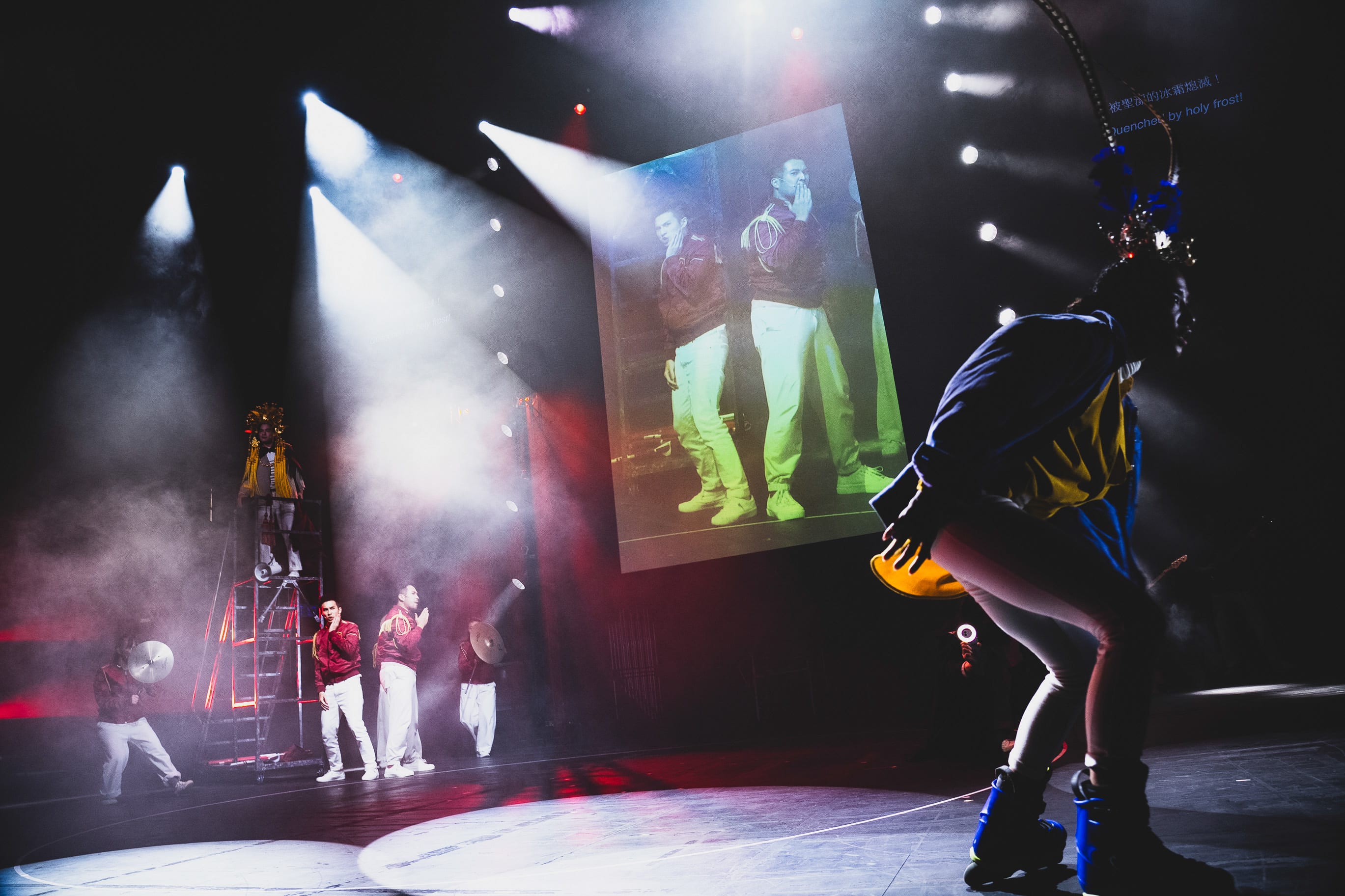 A dynamic stage shot from Rooftop Productions' contemporary theatre performance, 'Songs of Innocence and Experience' (WestK). In the foreground, a performer in blue rollerblades and a yellow jacket leans forward, capturing the energetic movement. On the left, actors including Cheung Cheuk-hang, Mok Kok-pong, and Caroline Chan are positioned on and around a metal stepladder and the stage floor. A large screen displays a projected image of the performers in red jackets, creating a multi-layered visual effect enhanced by dramatic stage lighting and hazy fog. 'Songs of Innocence and Experience (WestK).'