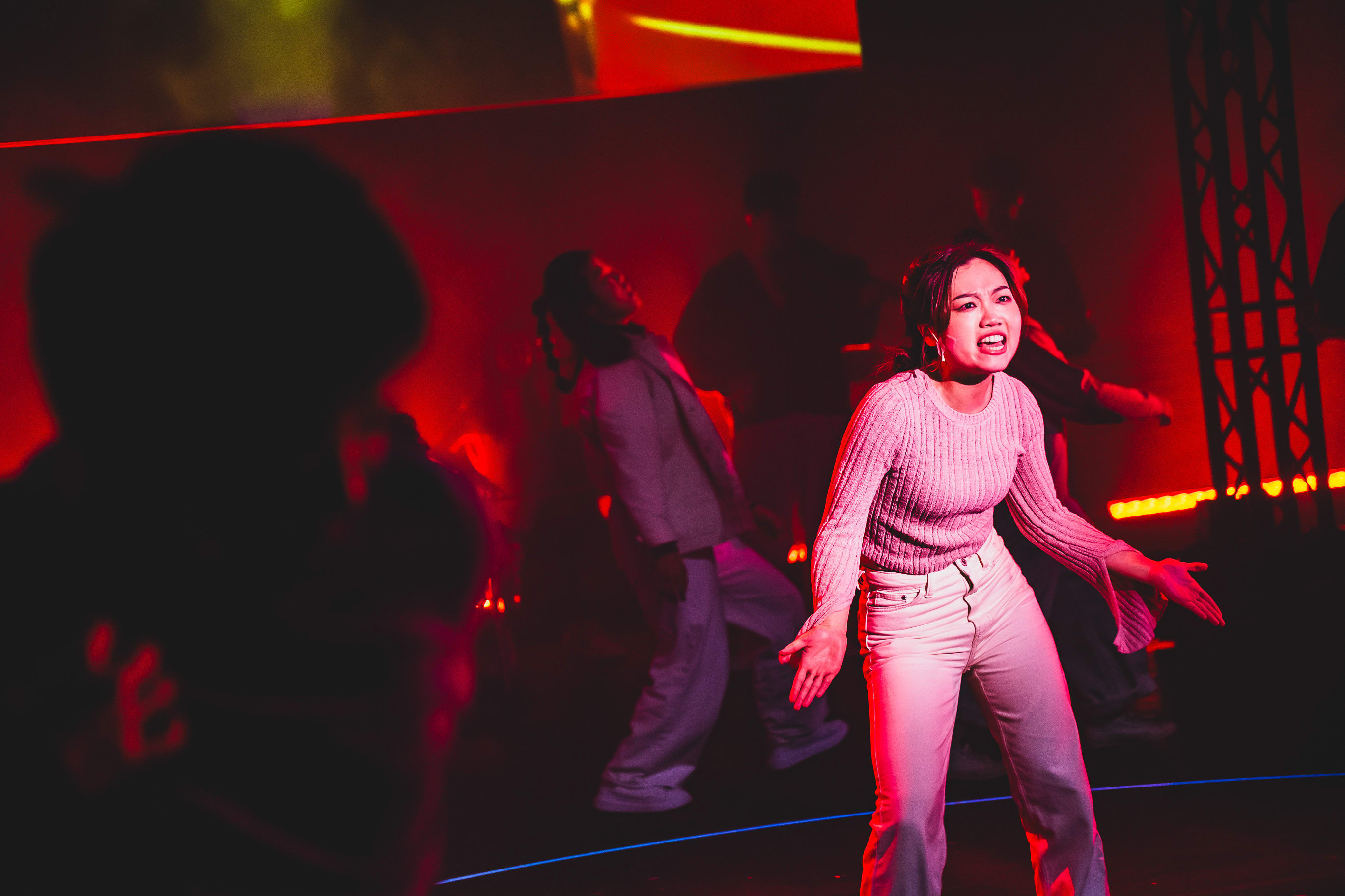 In a dramatic scene from Rooftop Productions' contemporary theatre piece 'Songs of Innocence and Experience' at WestK, actor Lee On-sang (as Moloch) stands center stage bathed in intense red lighting. She appears to be singing or delivering an emotional line with an intense, expressive gaze. Behind her, another performer in white moves across the dimly lit, industrial-style stage. 'Songs of Innocence and Experience (WestK).'