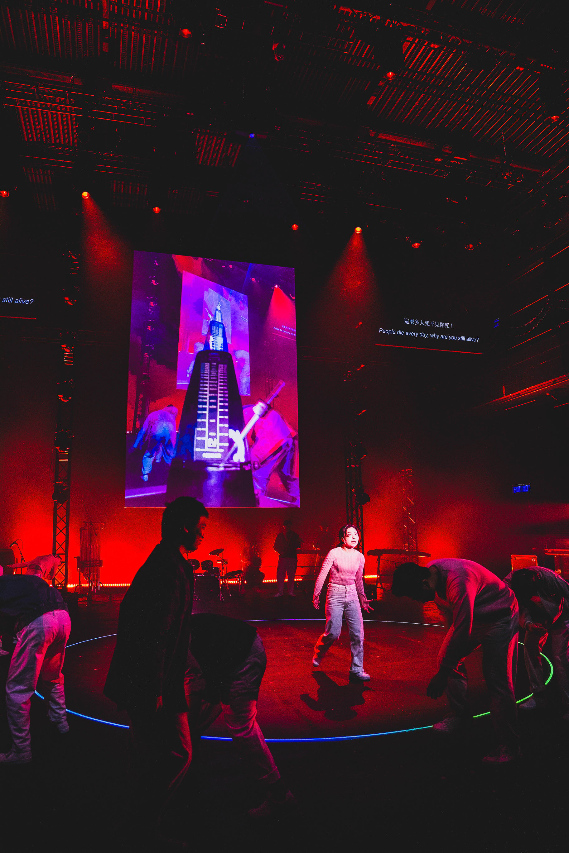 On a dimly lit stage bathed in intense red light, performers from Rooftop Productions engage in a powerful contemporary dance sequence during the Hong Kong theatrical production 'Songs of Innocence and Experience' at WestK. In the center, Lee On-sang (as Moloch) stands out as a woman in a light-colored top and jeans, surrounded by figures leaning forward in a circular formation, while a giant screen behind them projects an image of a metronome. Reflecting on the show's dark themes, the performers evoke the haunting question: 'People die every day, why are you still alive?'