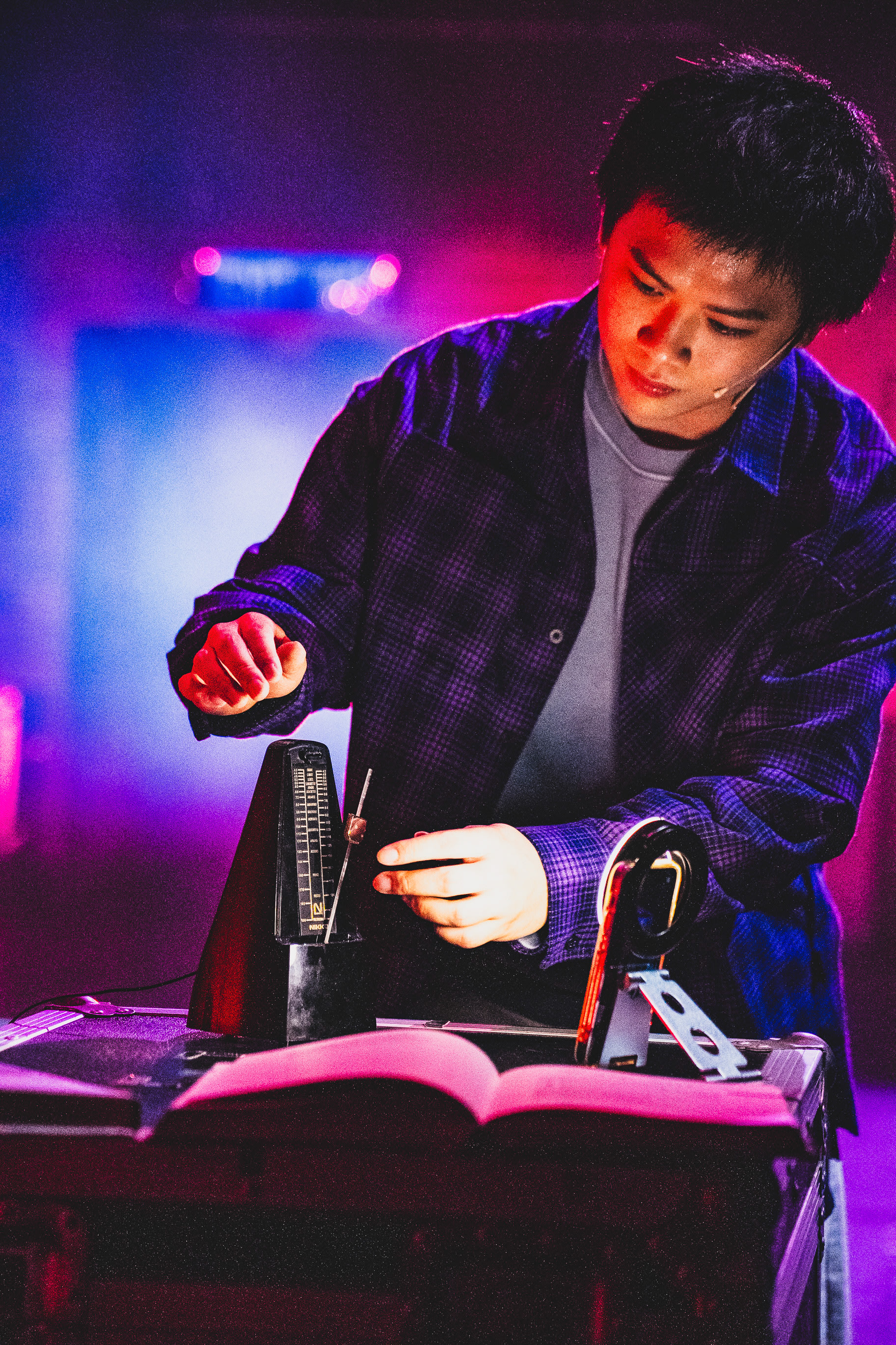 In a dramatic scene from Rooftop Productions' contemporary theatre piece 'Songs of Innocence and Experience' at WestK, actor Chew Yu-yeung, portraying the character Mammon, is captured under moody blue and purple stage lighting. He is intensely focused, adjusting the weight on a mechanical metronome sitting on a dark stage case, with an open script or book resting in front of him. The atmosphere reflects the experimental aesthetic of Hong Kong independent theatre. 'Songs of Innocence and Experience (WestK)'
