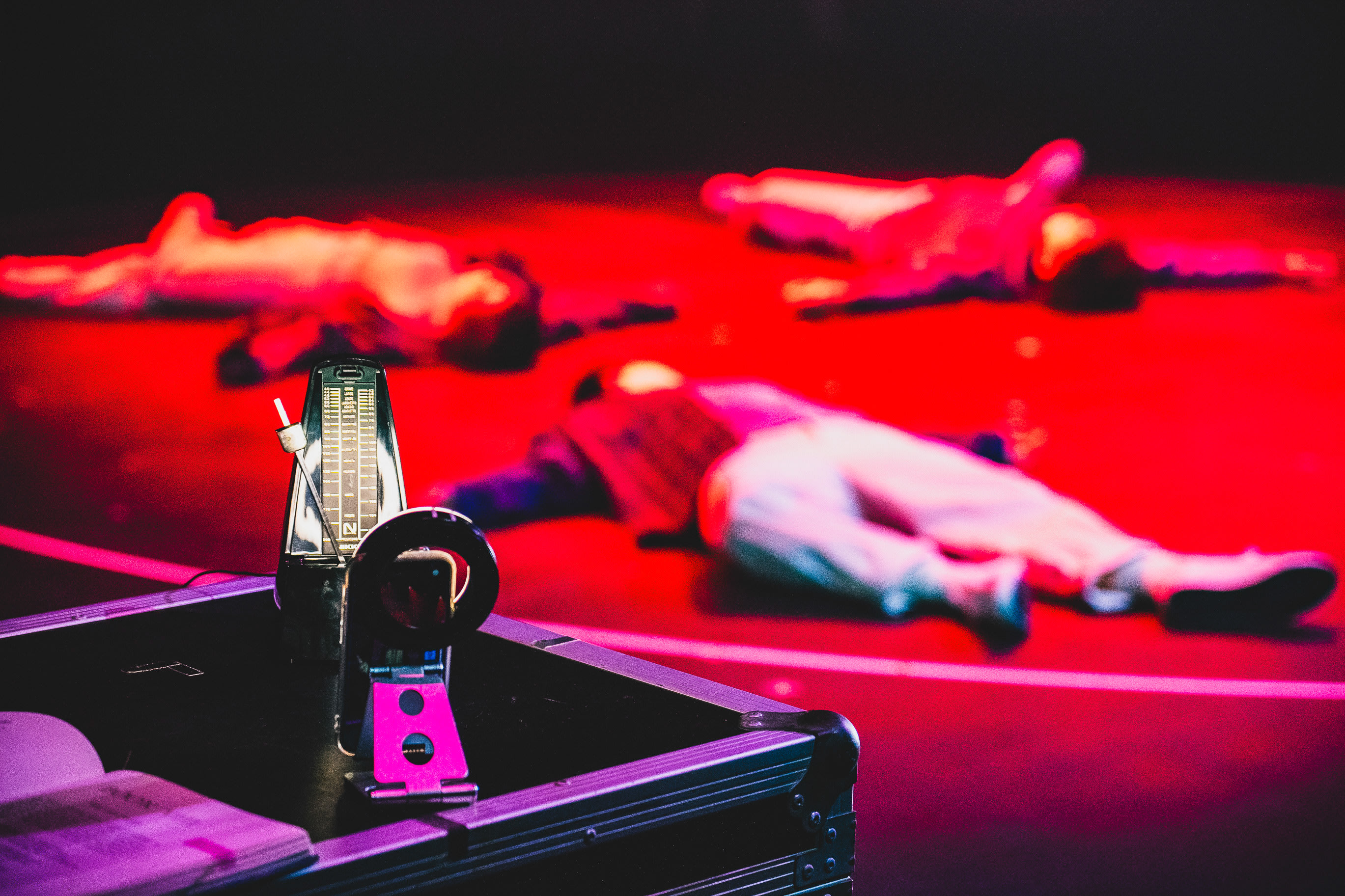 A dramatic stage shot from Rooftop Productions’ contemporary theatre piece 'Songs of Innocence and Experience' (WestK). In the foreground, a metronome and a small camera rest on a equipment case, while three performers lie prone in stillness on a stage bathed in intense red light, creating an atmosphere of vulnerability and tension. Songs of Innocence and Experience (WestK)