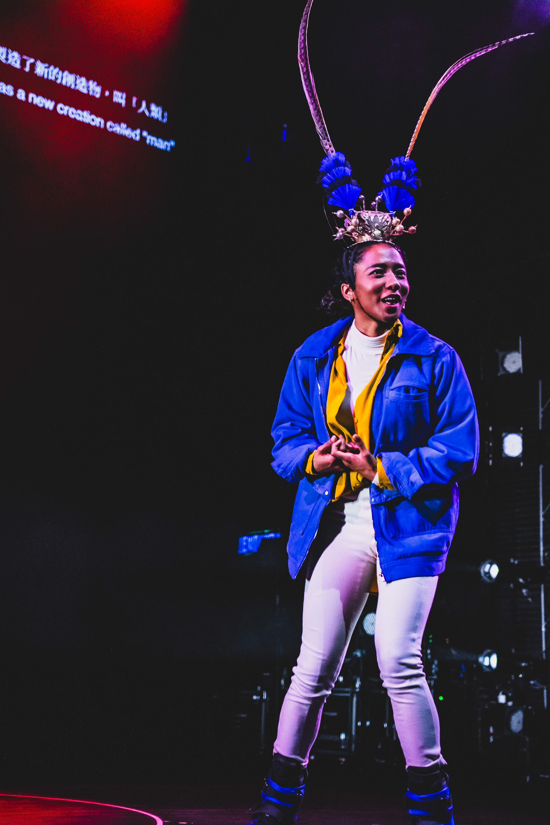 Caroline Chan performs on stage in Rooftop Productions' contemporary theatre piece, Songs of Innocence and Experience. Wearing a modern outfit paired with an elaborate, traditional-inspired headpiece with long, curved antennae, she gazes upward with an expressive, storytelling gesture under dramatic stage lighting. On the screen behind her, the text reads: "as a new creation called 'man'"