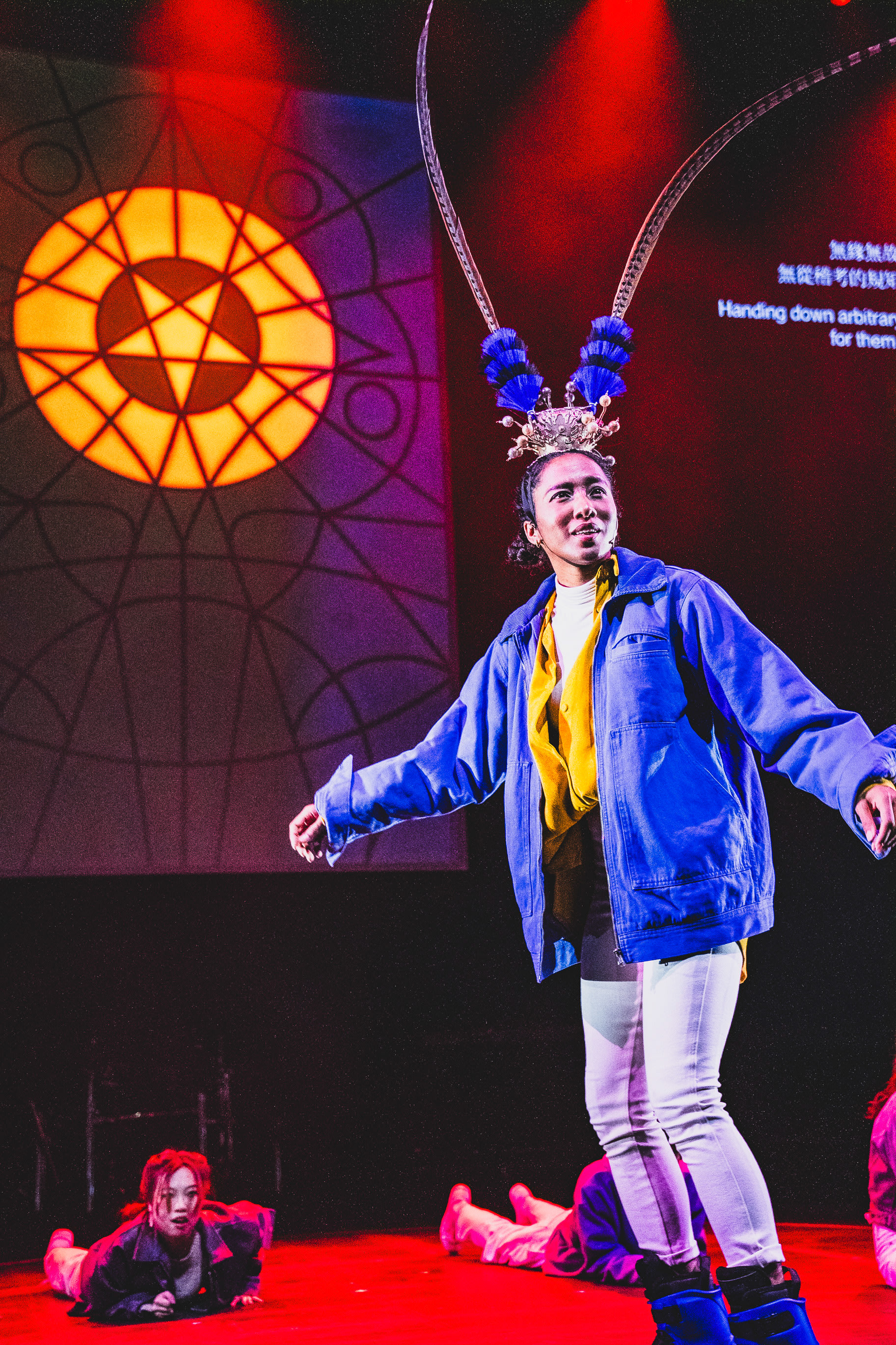 On a stage bathed in vibrant red lighting, performer Lee On-sang takes center stage in a contemporary production of Rooftop Productions' 'Songs of Innocence and Experience' at WestK. She is wearing a modern blue jacket and white trousers, adorned with a traditional opera-style headpiece featuring long, dramatic pheasant feathers. In the background, a large projection of a geometric pentagram pattern mirrors the theatrical intensity, while another actor, Caroline Chan, is seen crouching on the floor in the lower left. 'Songs of Innocence and Experience (WestK)'