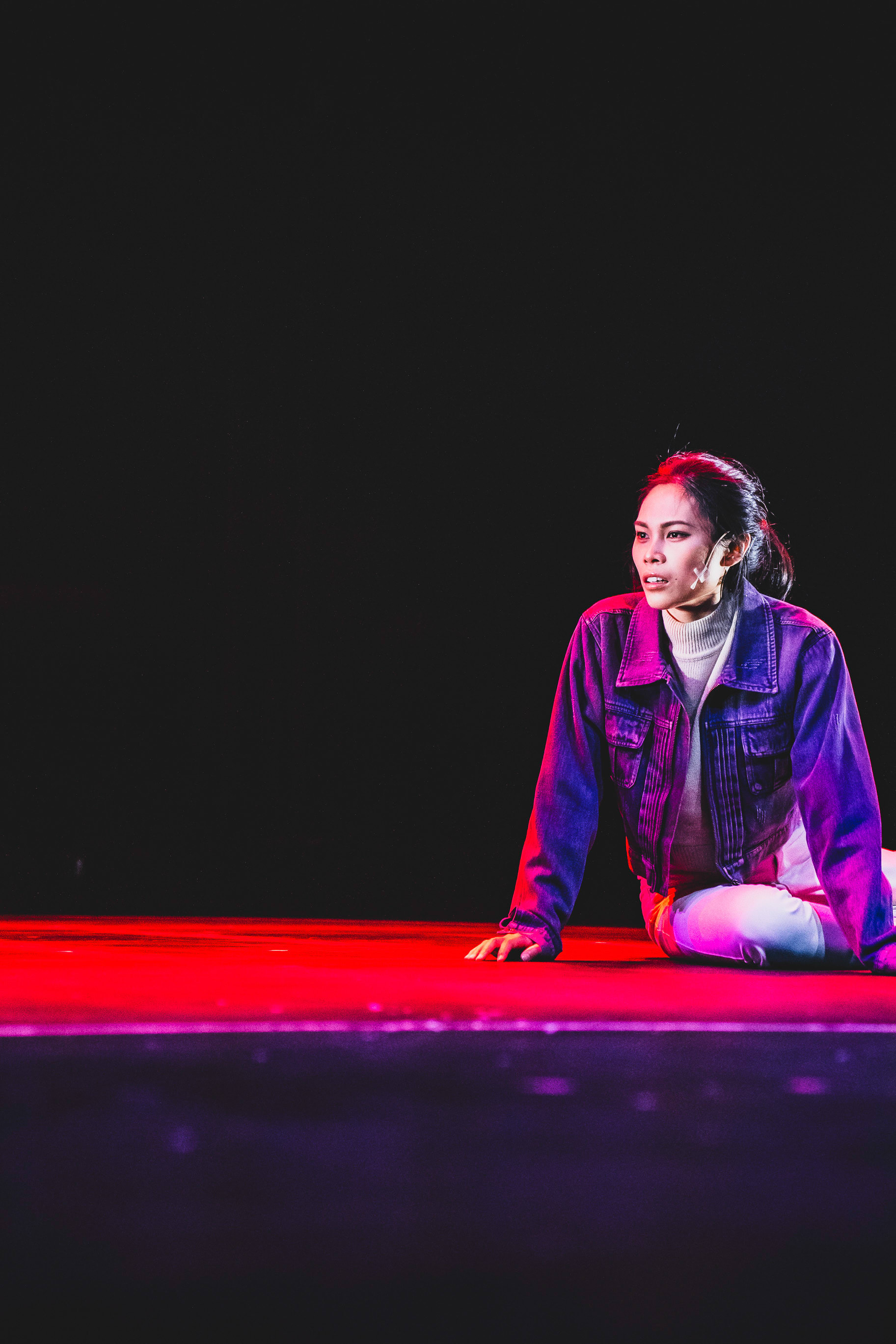 A dramatic stage shot of performer Chin Wing-kar (portraying Belial) during a performance of the contemporary Hong Kong theatre production 'Songs of Innocence and Experience' at the West Kowloon Cultural District (WestK). She is sitting on a stage floor bathed in vivid red and purple lighting, wearing a denim jacket and light-colored turtleneck, looking off-stage with a focused and emotional expression. This scene is from 'Songs of Innocence and Experience' (WestK).