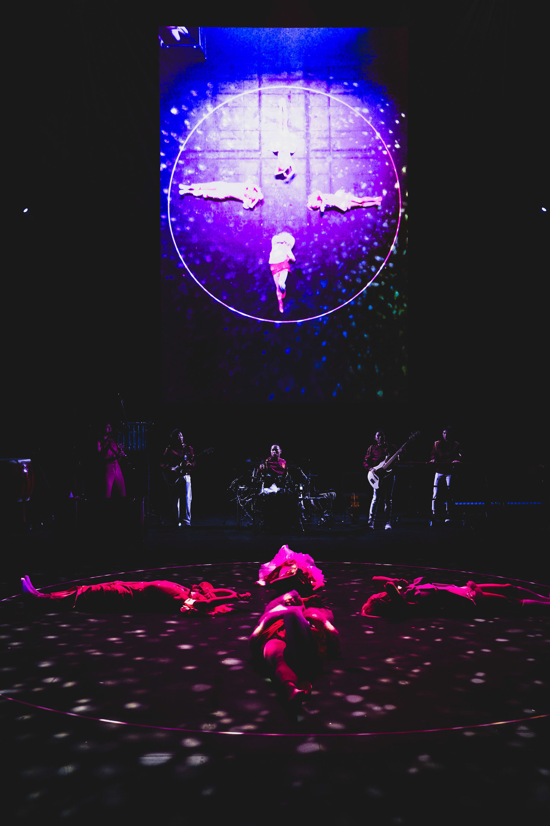 On a dark circular stage, four performers lie in a star formation, illuminated by dramatic pools of purple and red light. Behind them, a large vertical screen displays a mirrored projection of the same choreography, creating an immersive, multi-layered visual experience characteristic of contemporary Hong Kong theatre. This evocative scene is from the Rooftop Productions (天台製作) performance, 'Songs of Innocence and Experience' (WestK).