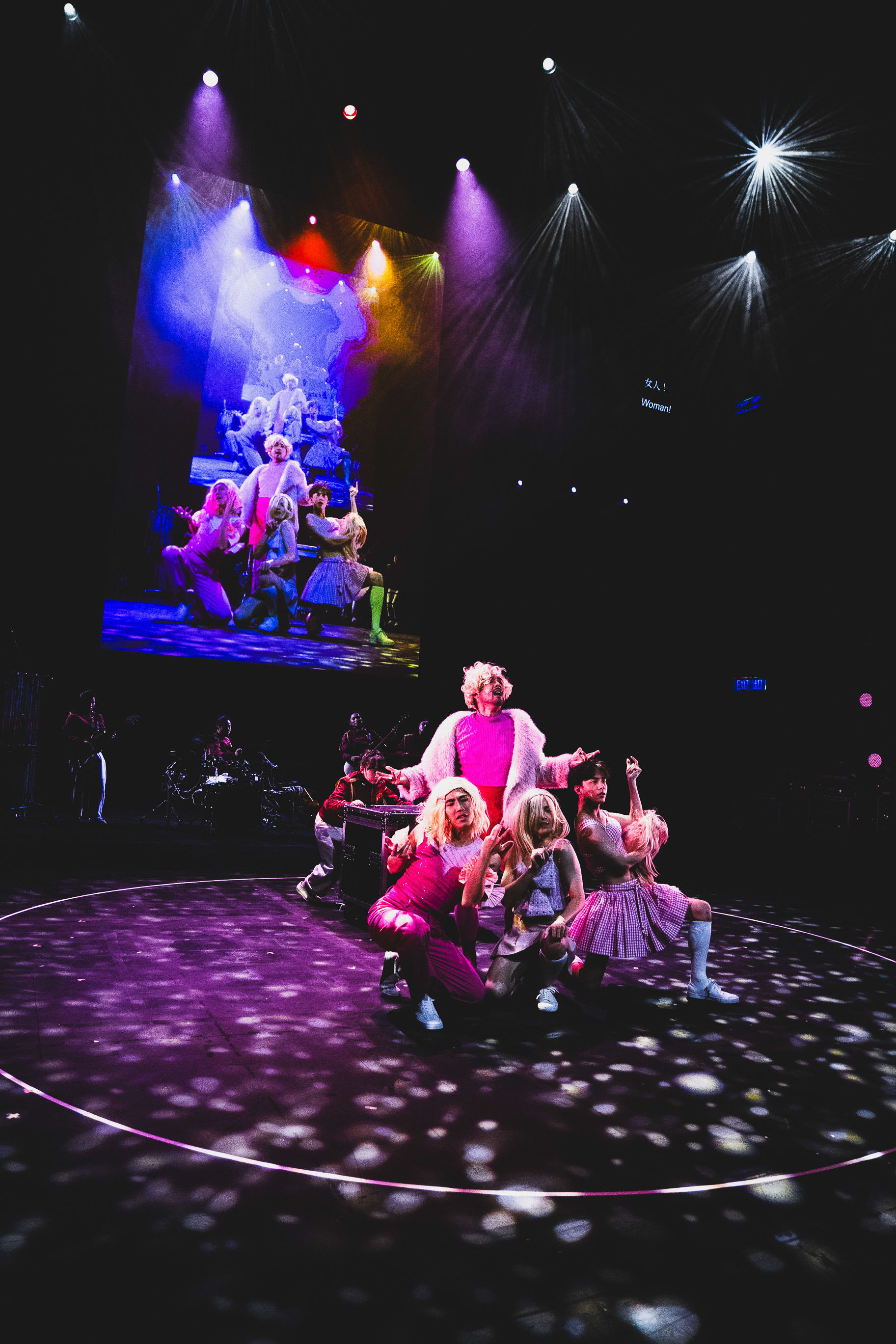 A wide shot of a dynamic performance of 'Songs of Innocence and Experience' at the West Kowloon Cultural District (WestK). On a circular, spotlighted stage, Beelzebub stands center stage wearing a white fur coat over a pink top, surrounded by other performers including an Intern Angel, all illuminated by vibrant, theatrical stage lighting. The backdrop features a large screen displaying imagery of the troupe, capturing the bold, avant-garde aesthetic of Rooftop Productions. 'Songs of Innocence and Experience (WestK)'