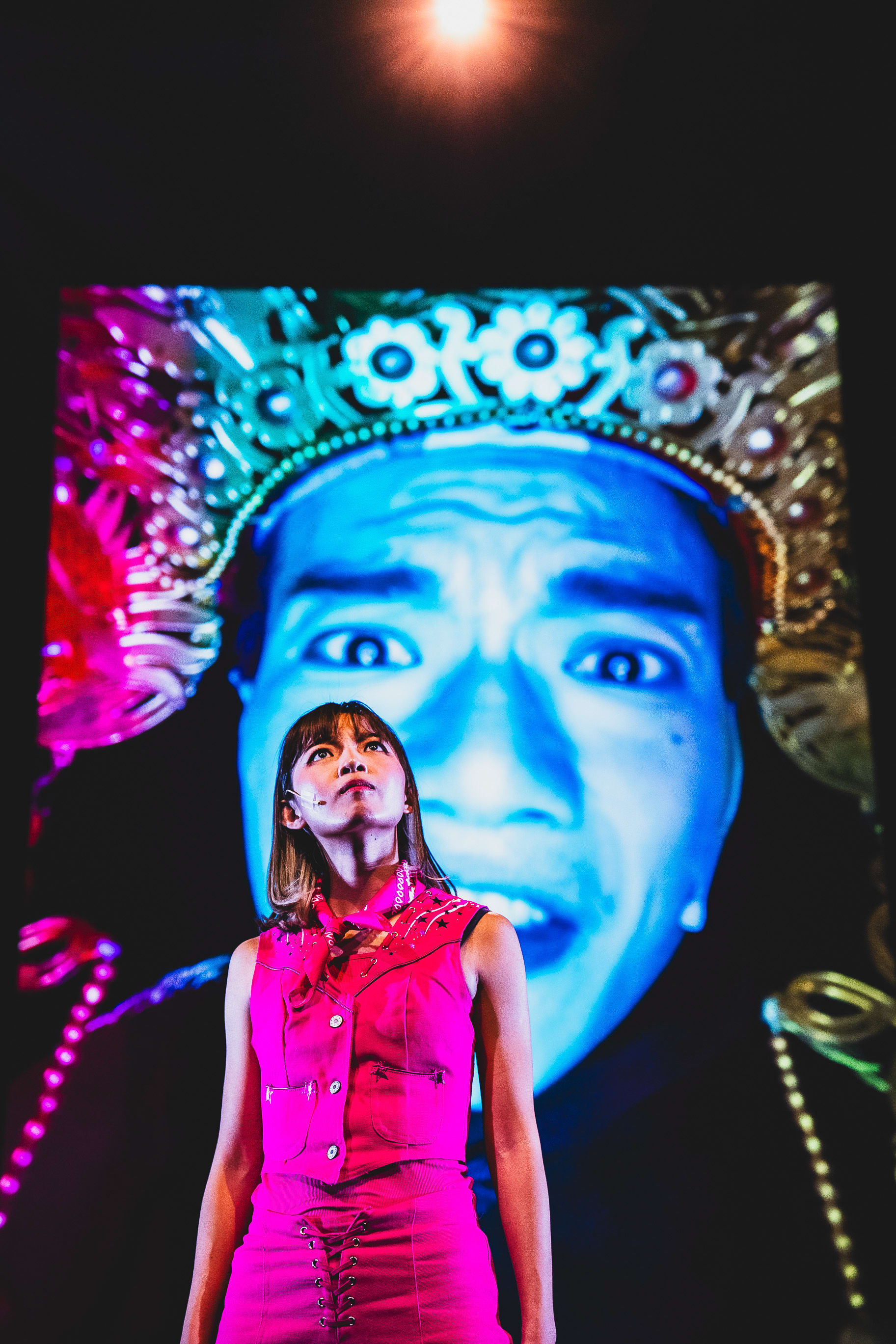 In a striking scene from the contemporary Hong Kong theatre production 'Songs of Innocence and Experience' (《天真與世故之歌》) at WestK, actor Sin Lok-yan stands center stage wearing a bright pink sleeveless vest and skirt. She gazes upward with a determined expression, illuminated by dramatic stage lighting. Behind her, a massive, high-contrast digital projection of Lo Man-chak in elaborate, traditional-style headgear looms over the stage, adding an intense and surreal layer to the performance. Songs of Innocence and Experience (WestK)