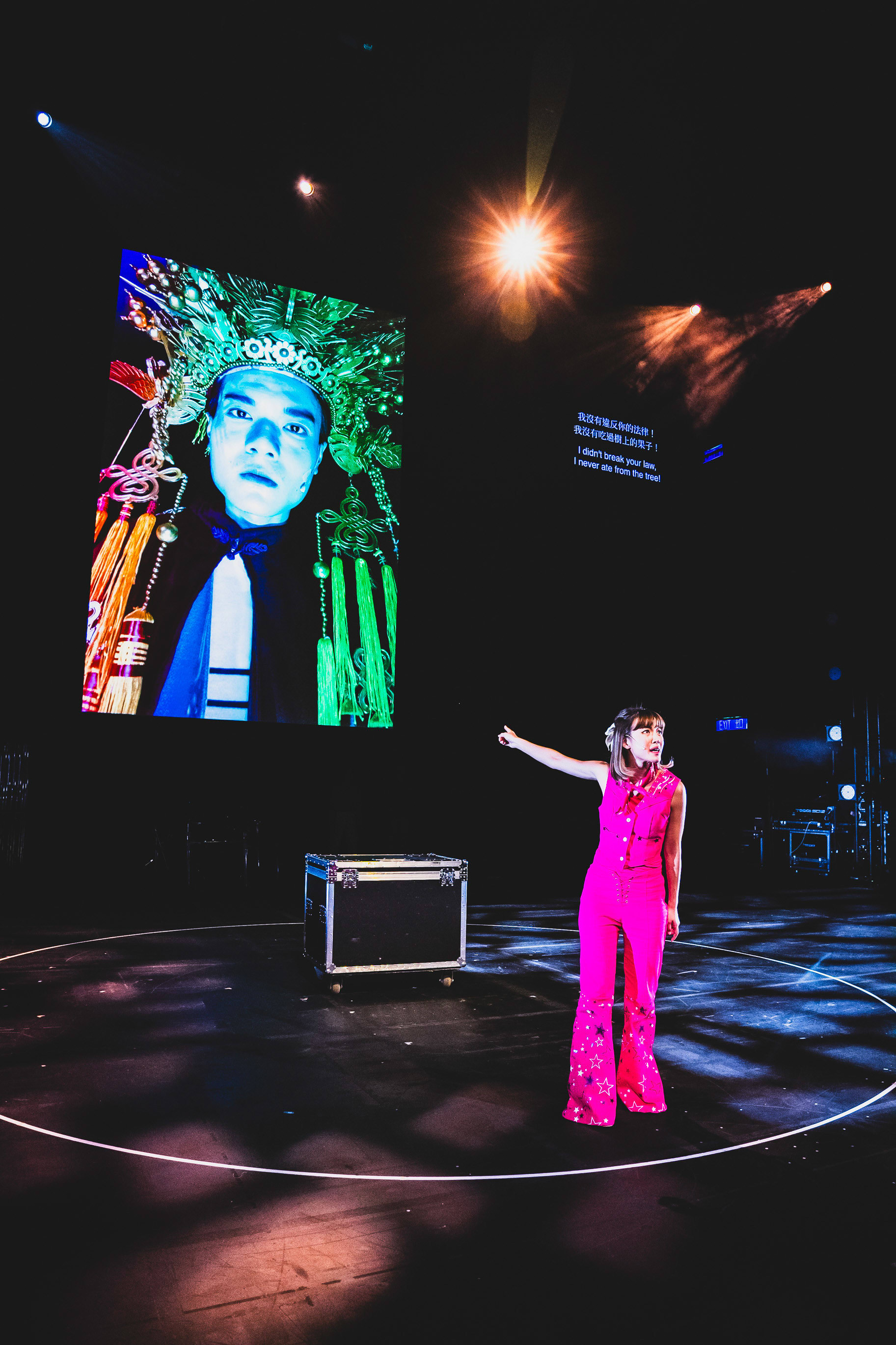 In this scene from Rooftop Productions’ contemporary theatre piece Songs of Innocence and Experience, actor Sin Lok-yan stands center stage in a bright pink jumpsuit, gesturing toward a large projected image of Lo Man-chak appearing as God. The stark, dramatic stage lighting highlights the interaction between the performer and the screen, reflecting themes of divinity and human innocence. As she points, Sin Lok-yan delivers the line: "I didn't break your law. I never ate from the tree."