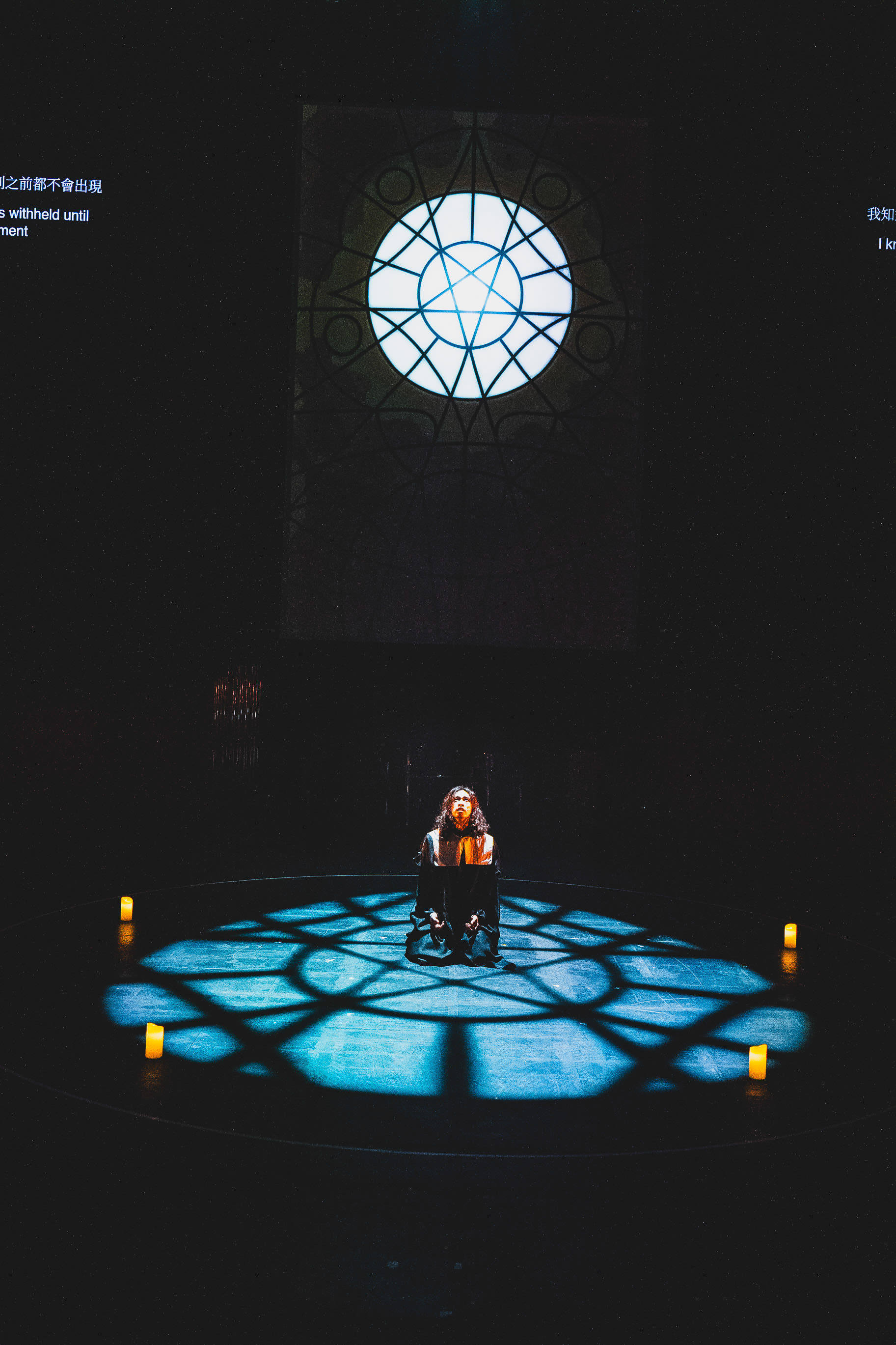A dramatic stage shot from Rooftop Productions' contemporary theatre performance, 'Songs of Innocence and Experience'. Actor Mak Ho-tin is centered on a dark circular stage, kneeling in a pool of blue, stained-glass-patterned light that projects from a luminous circular emblem above. The atmosphere is stark and introspective, capturing the performer in a moment of intense solitude. 'Milton in Jail'