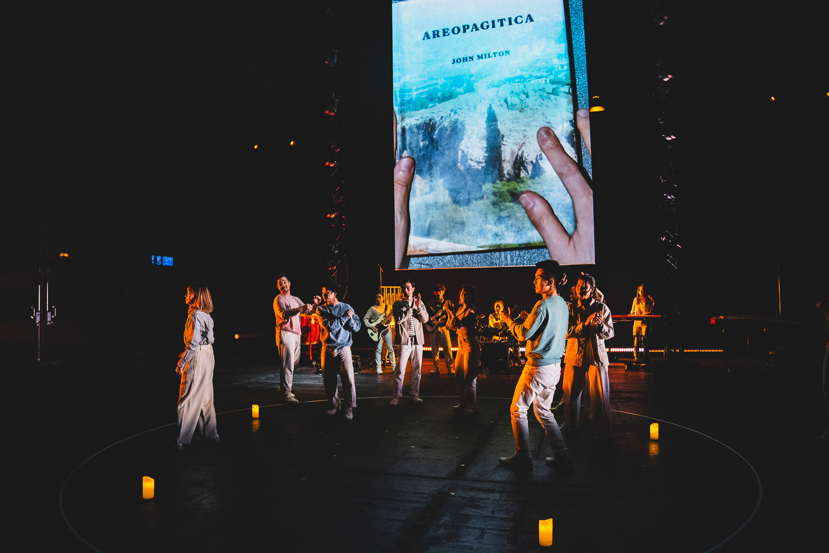 天台製作（Rooftop Productions）當代劇場演出《天真與世故之歌》（Songs of Innocence and Experience）（西九版）的舞台實景。舞台燈光昏暗，演員包括錢穎嘉、趙汝陽等表演者以圓形排列，地面上散佈著溫暖的蠟燭，營造出神秘而沉思的氛圍。舞台背景是一面巨大的螢幕，展示著雙手捧著約翰·彌爾頓的《Areopagitica》的影像。這場跨界藝術表演展現了香港當代劇場的深度。演員在台上唸道：「Areopagitica。」
