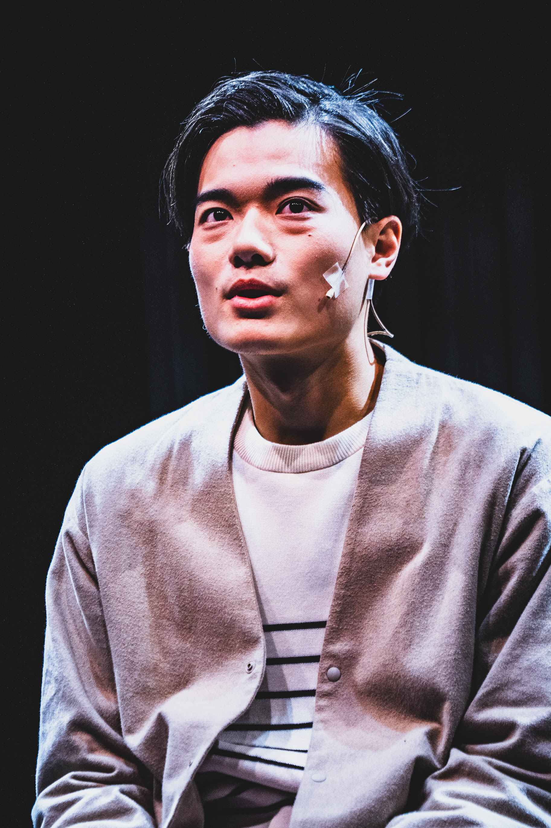 Close-up of actor Lo Man-chak performing in Rooftop Productions' contemporary theatre piece 'Songs of Innocence and Experience' at West Kowloon. The actor, portraying the role of Himself/God, is captured under dramatic stage lighting with a headset microphone visible, his expression contemplative and searching. 'Are you happy?'
