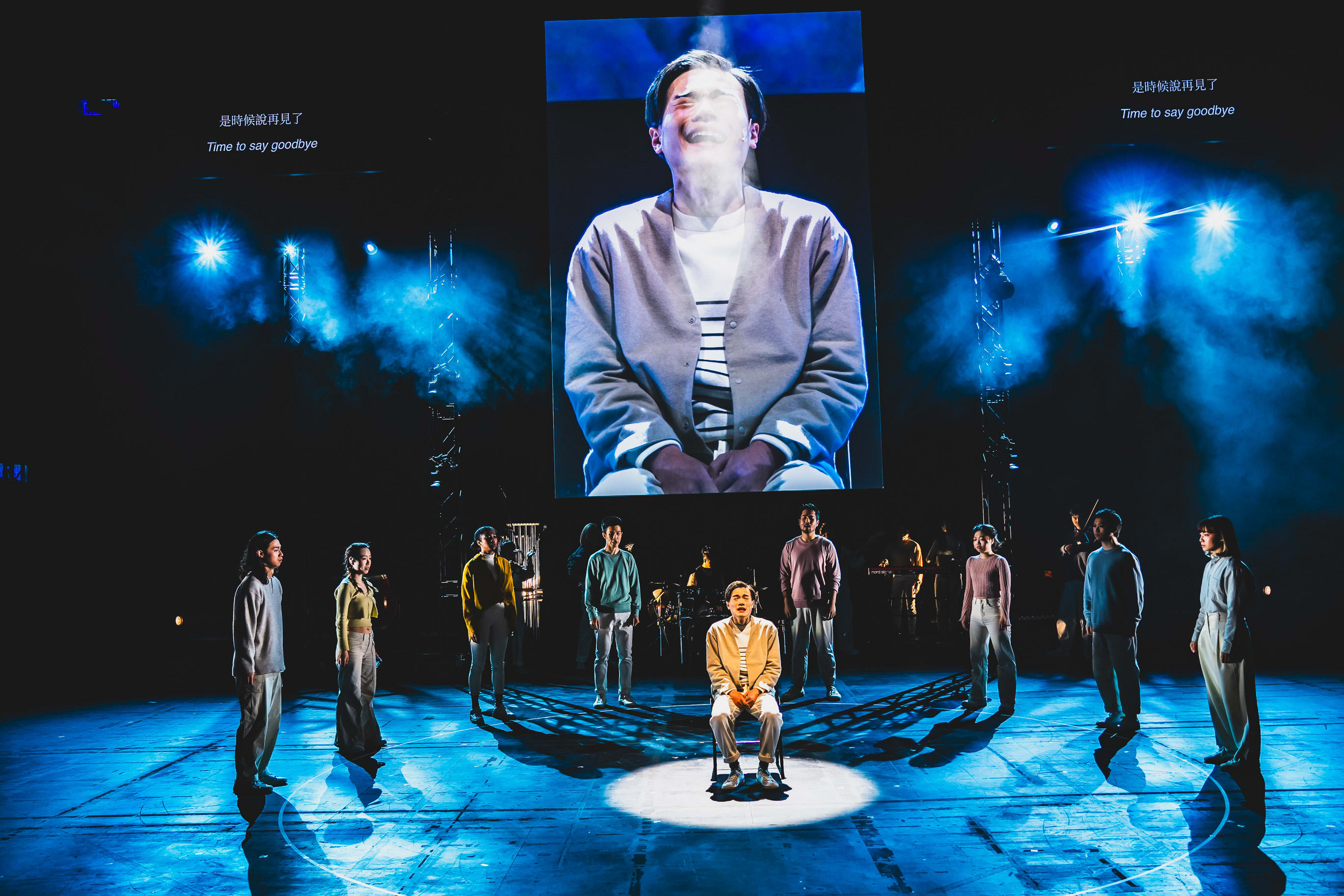 天台製作（Rooftop Productions）於西九文化區上演的當代劇場作品《天真與世故之歌》（Songs of Innocence and Experience）。舞台燈光冷冽，背景投影著一位神情痛苦的男子巨大特寫，螢幕上寫著「Time to say goodbye」。舞台上，演員們分散站立，營造出深沉的戲劇氛圍；飾演密爾頓（Milton）的麥晧鈿獨自坐在舞台中央的聚光燈下。劇中角色唱道：「Cause now the guilt is all mine」
